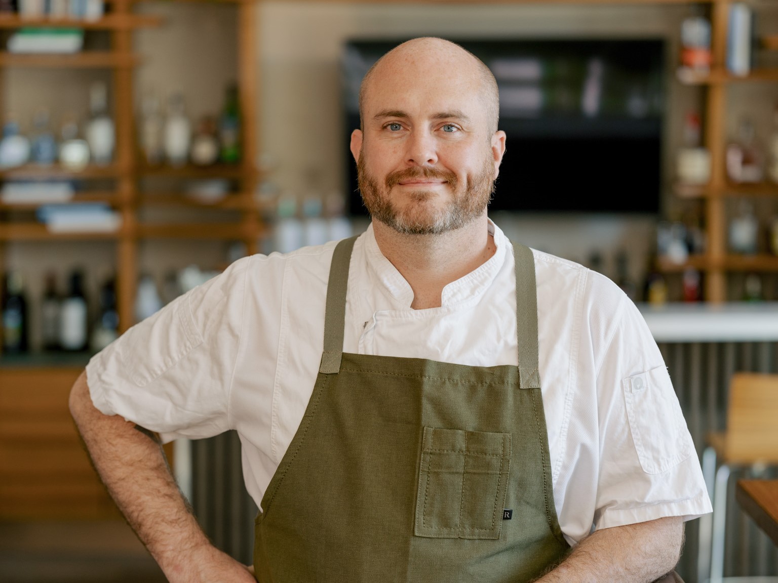 A bald chef with a beard, smiling in an oceanfront restaurant, wearing a white shirt and green apron, with shelves of bottles in the background.