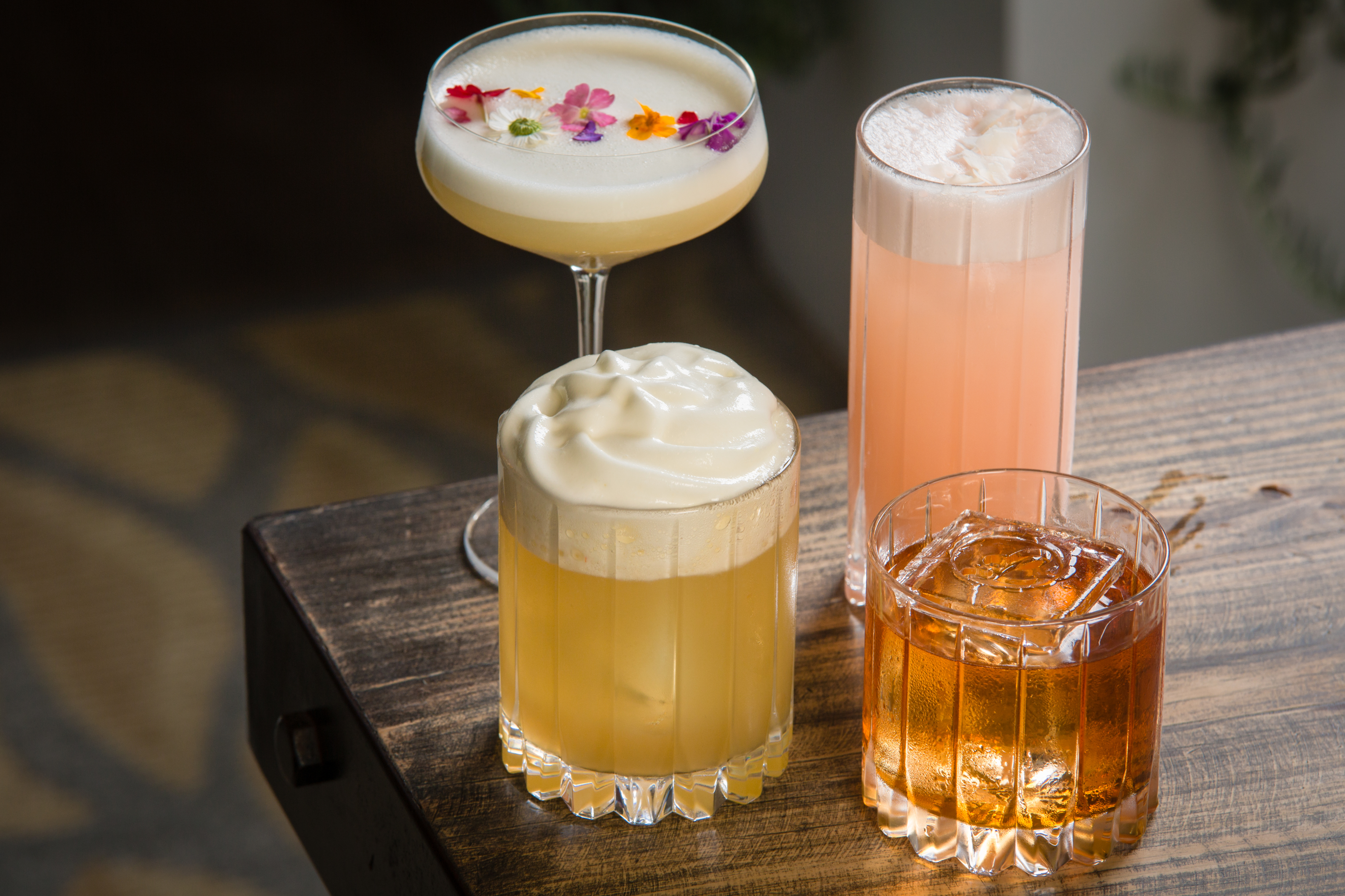 Four different cocktails on a wooden table at an open-air patio, each with distinct presentations ranging from floral garnishes to frothy toppings.