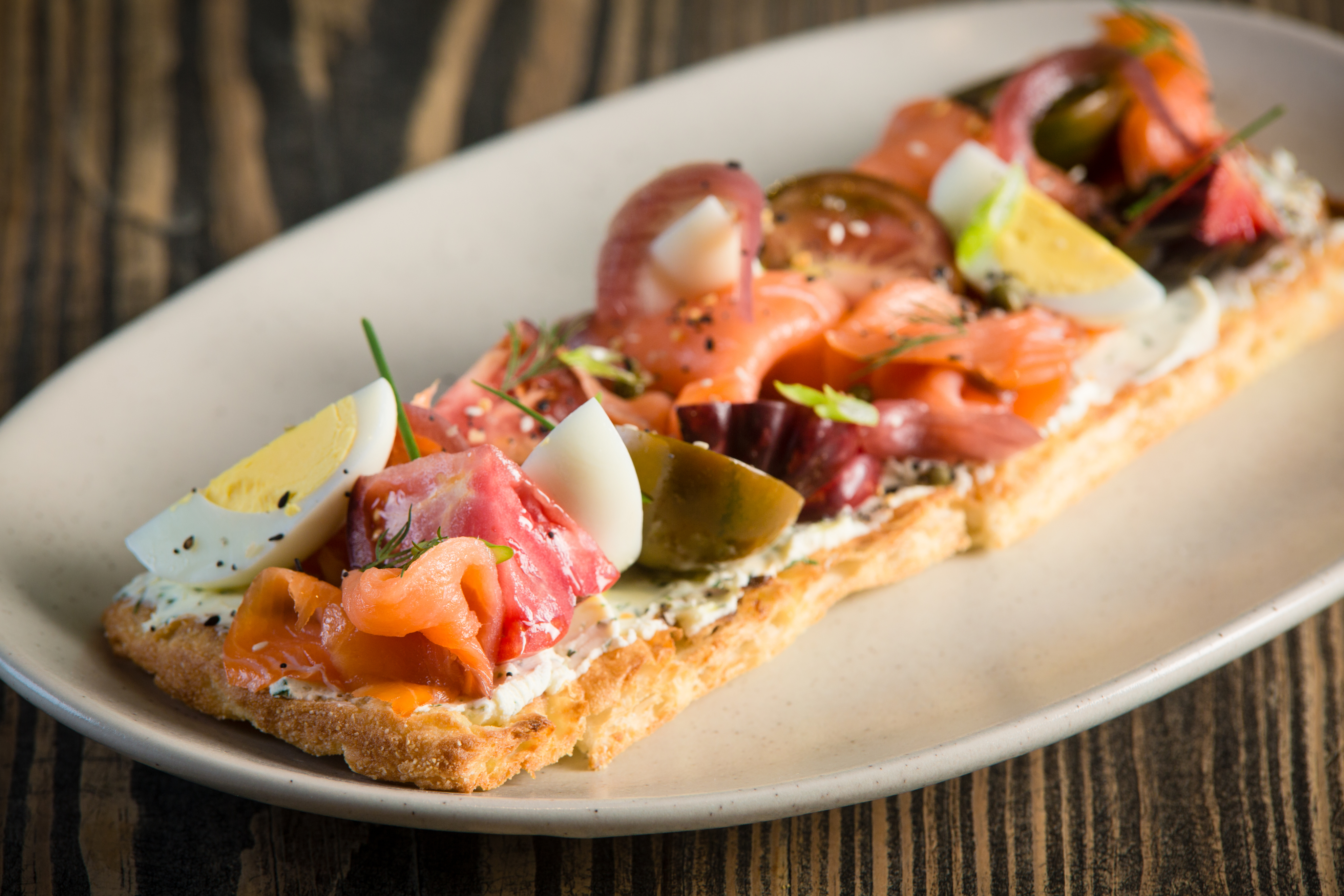 An oval plate holding a lavish open-faced sandwich topped with smoked salmon, sliced boiled eggs, tomatoes, red onions, and fresh herbs from locally-sourced food, presented on a rustic wooden table.