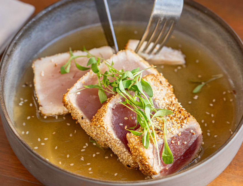 A bowl of seared sesame-crusted tuna slices served in broth, garnished with greens. Forks are holding one of the slices.