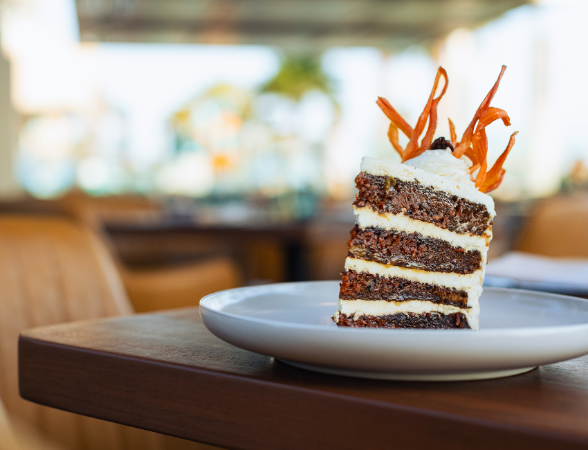 A slice of carrot cake with cream cheese frosting and decorative carrot strips graces a white plate, set on a wooden table in a cozy cafe setting. This delightful dessert is a must-try from our menu.