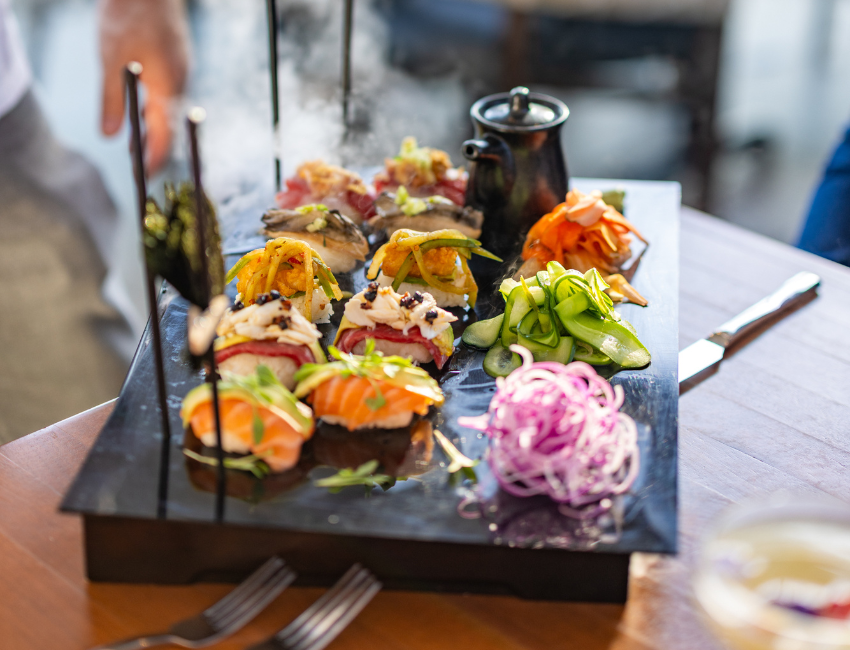 A black platter with assorted sushi, sliced vegetables, and a small black teapot, served on a wooden table with utensils nearby.