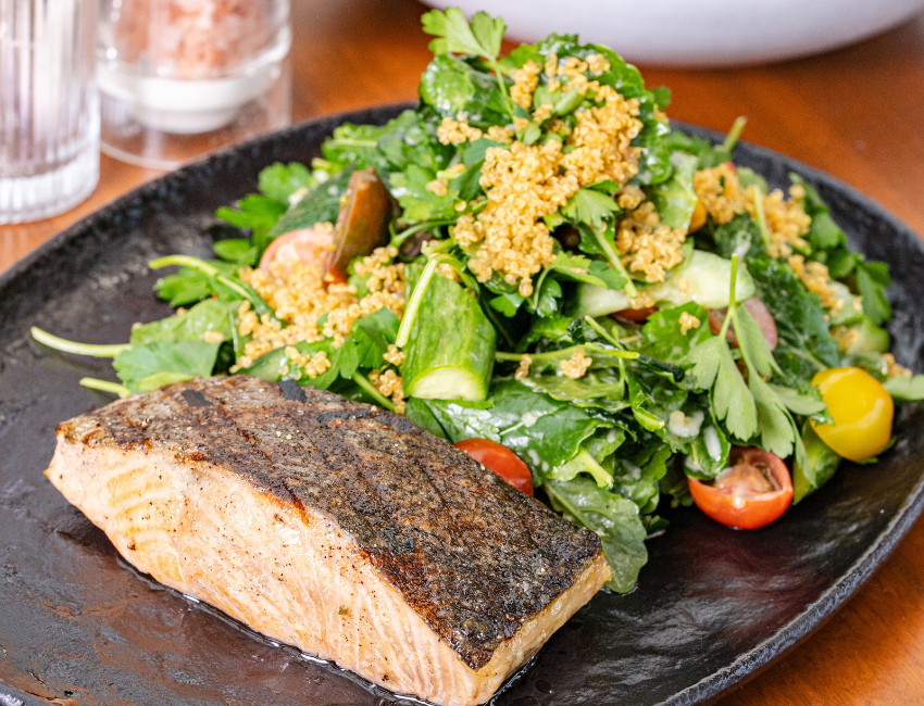 A cooked salmon fillet with crispy skin is served next to a fresh green salad with cherry tomatoes and cucumbers on a black plate.