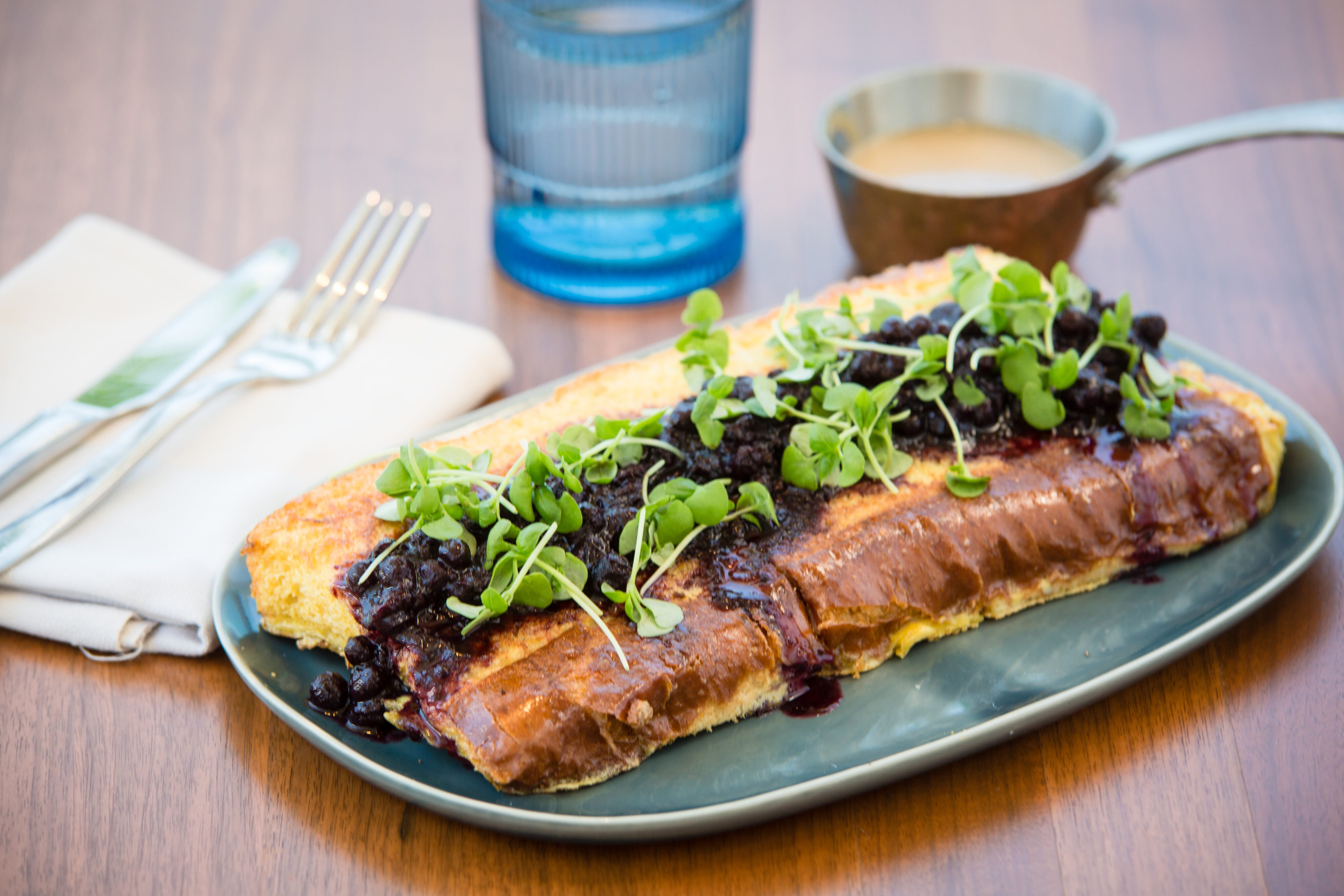 A plated dish featuring pork tenderloin topped with blueberry sauce and greens, perfect for a leisurely weekend brunch. Served on a rectangular dish with cutlery, a napkin, a blue glass, and a small saucepan of sauce on a wooden table.