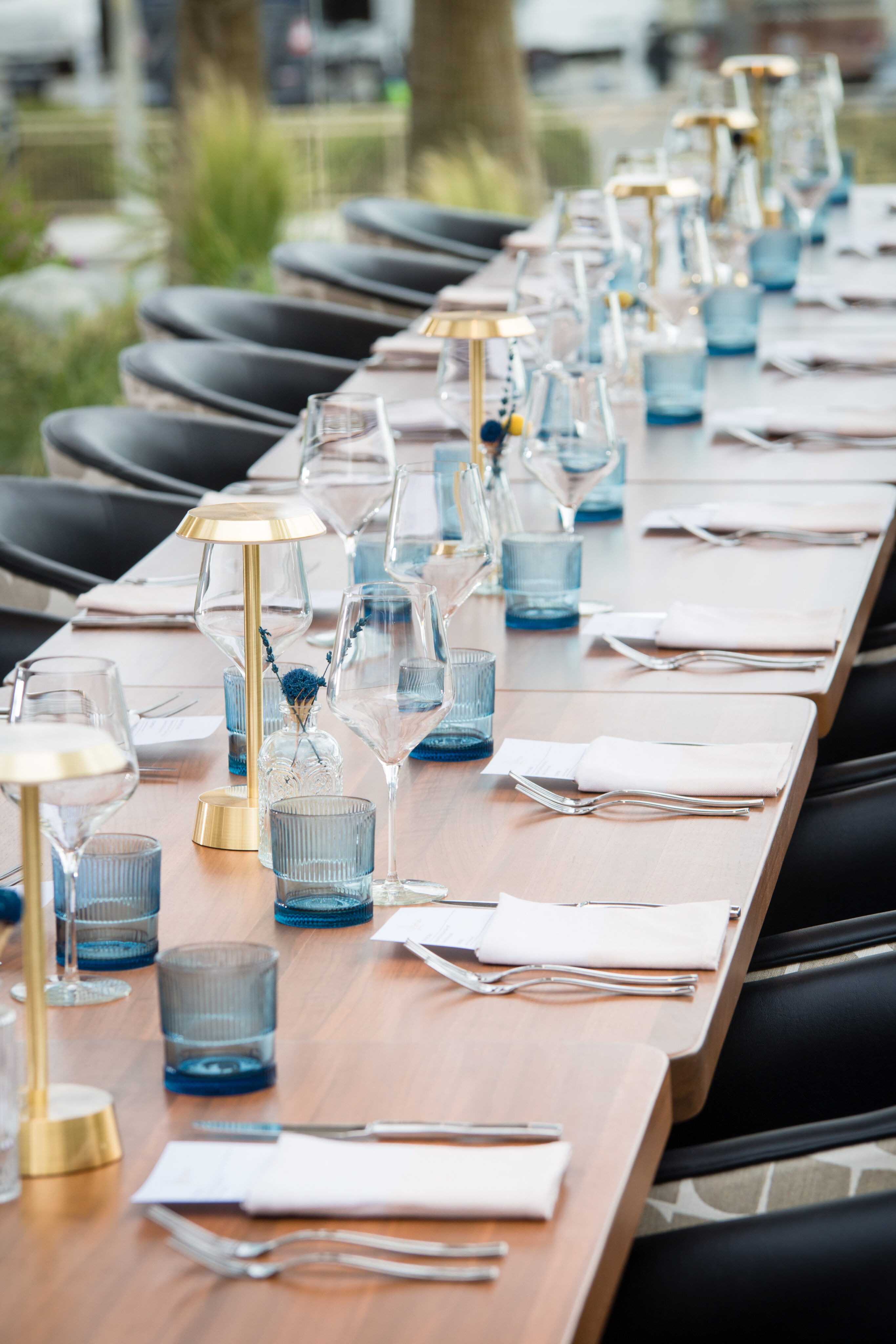 A long dining table is set with blue and clear glasses, gold and white lamps, white napkins, and cutlery. Black chairs are arranged along both sides of the table.