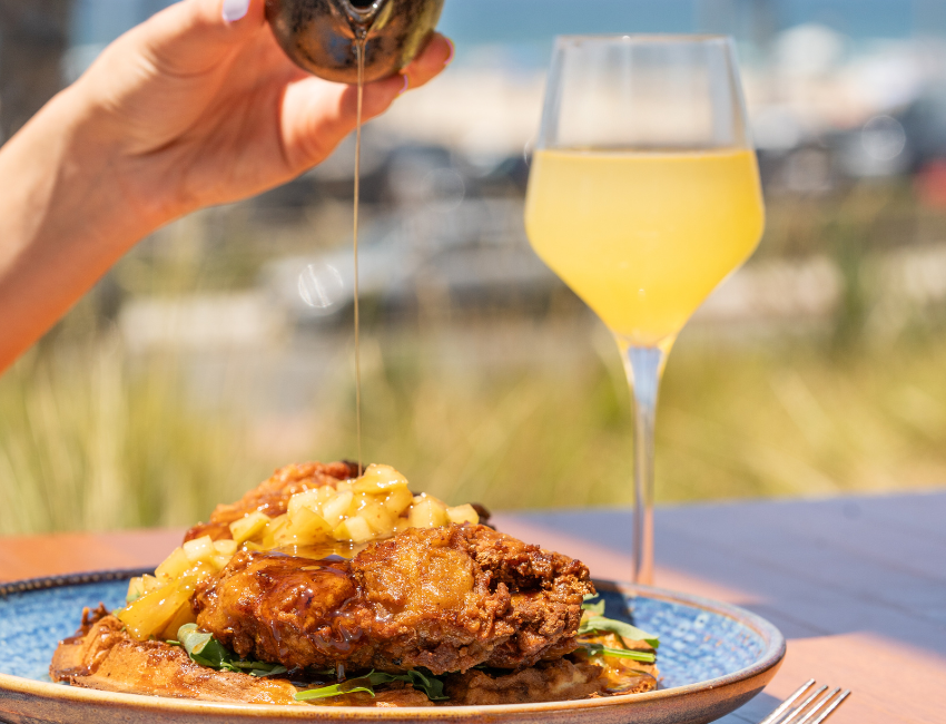 A hand pours syrup over fried chicken and waffles topped with diced fruit, perfect for a weekend brunch, with a glass of orange drink beside the plate on an outdoor table.