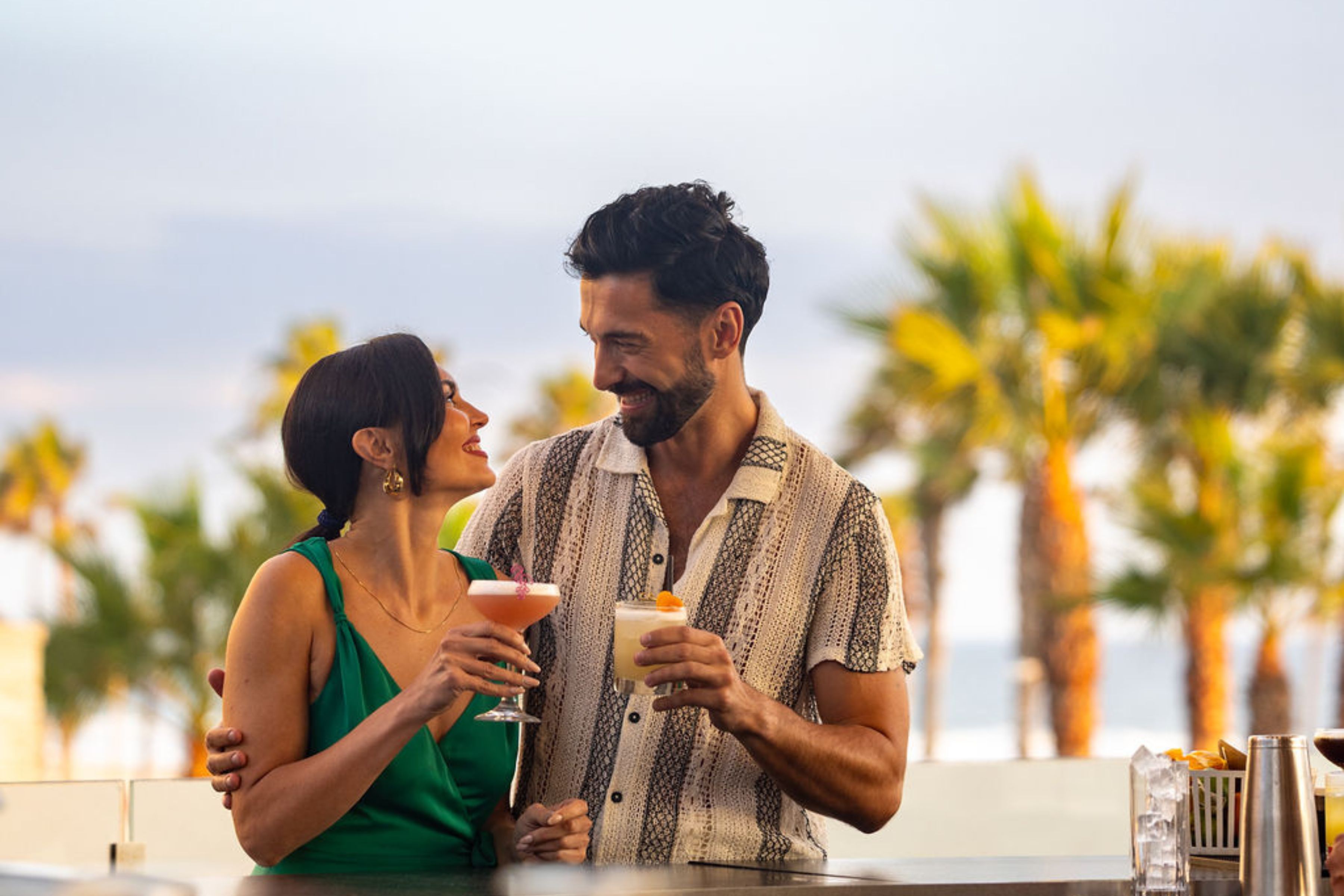 A man and woman stand close together outdoors at Happy Hour, smiling at each other while holding cocktails, with palm trees in the background.
