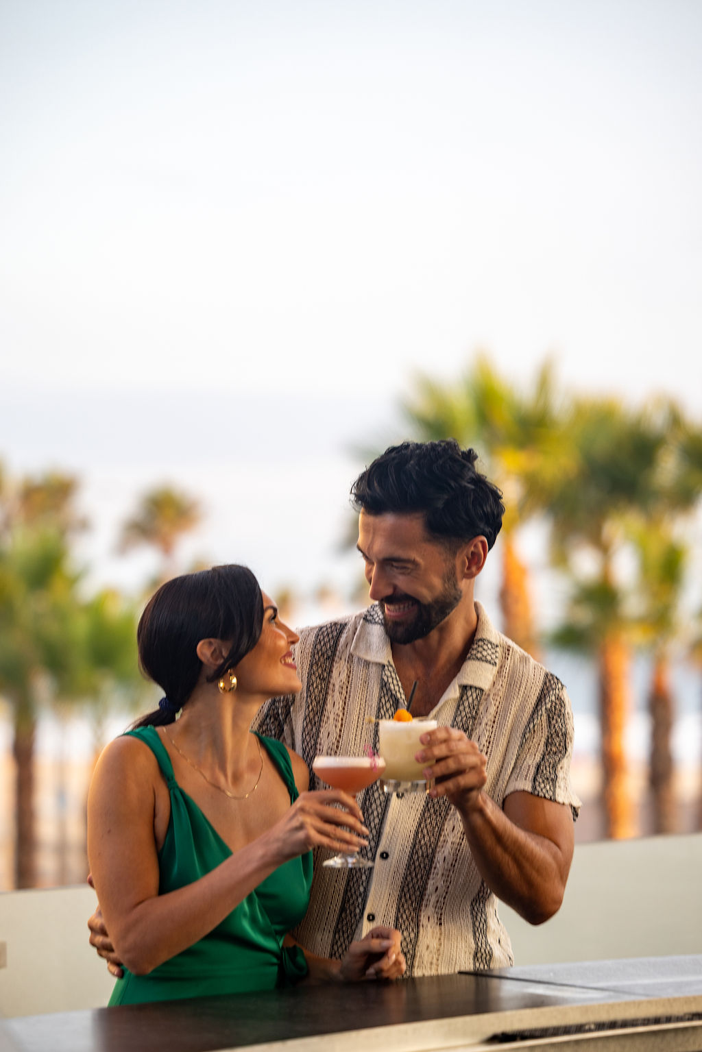 A man and woman stand outdoors, smiling at each other and holding cocktails during Happy Hour, with palm trees blurred in the background.