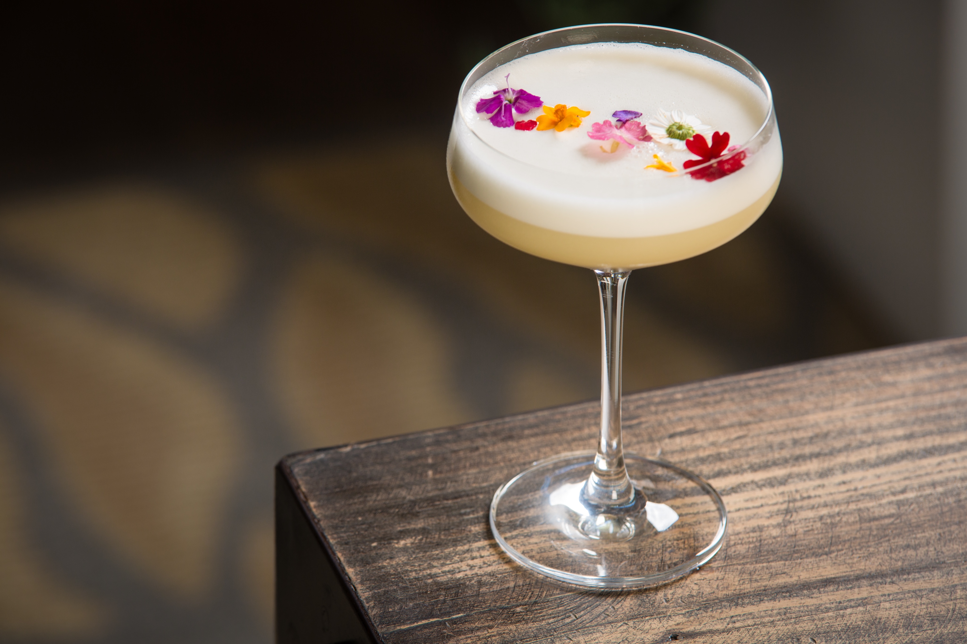 A coupe glass filled with a creamy white cocktail, garnished with colorful edible flowers, placed on a wooden surface - perfect for an enchanting treehouse happy hour.