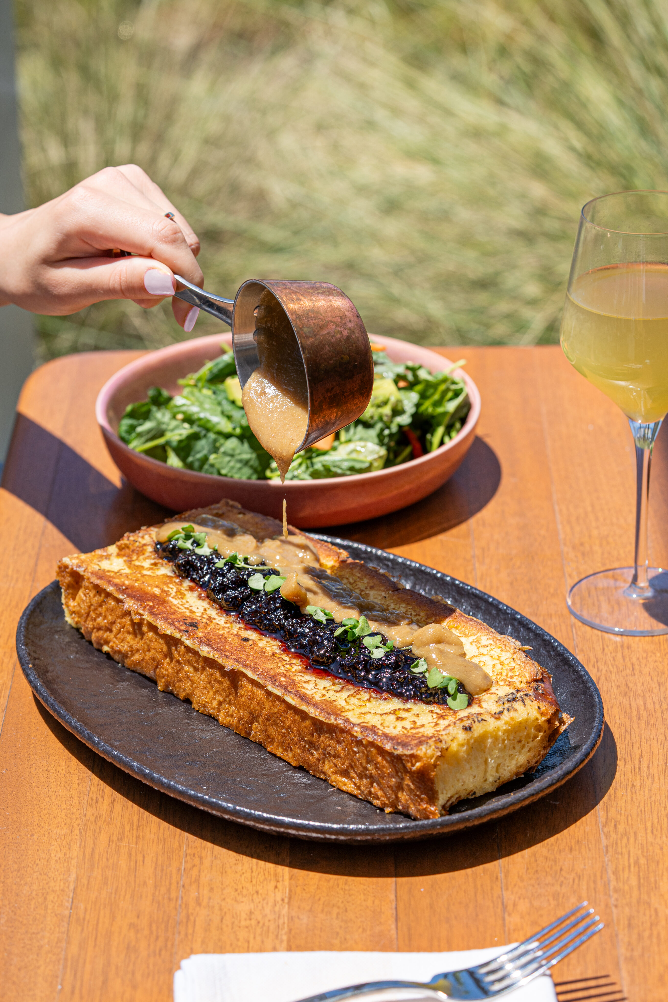 A hand pours sauce over a rectangular savory pastry topped with greens and a dark spread, served with salad and a glass of white wine—perfect for a relaxing Weekend Brunch at Lōrea on a wooden table.