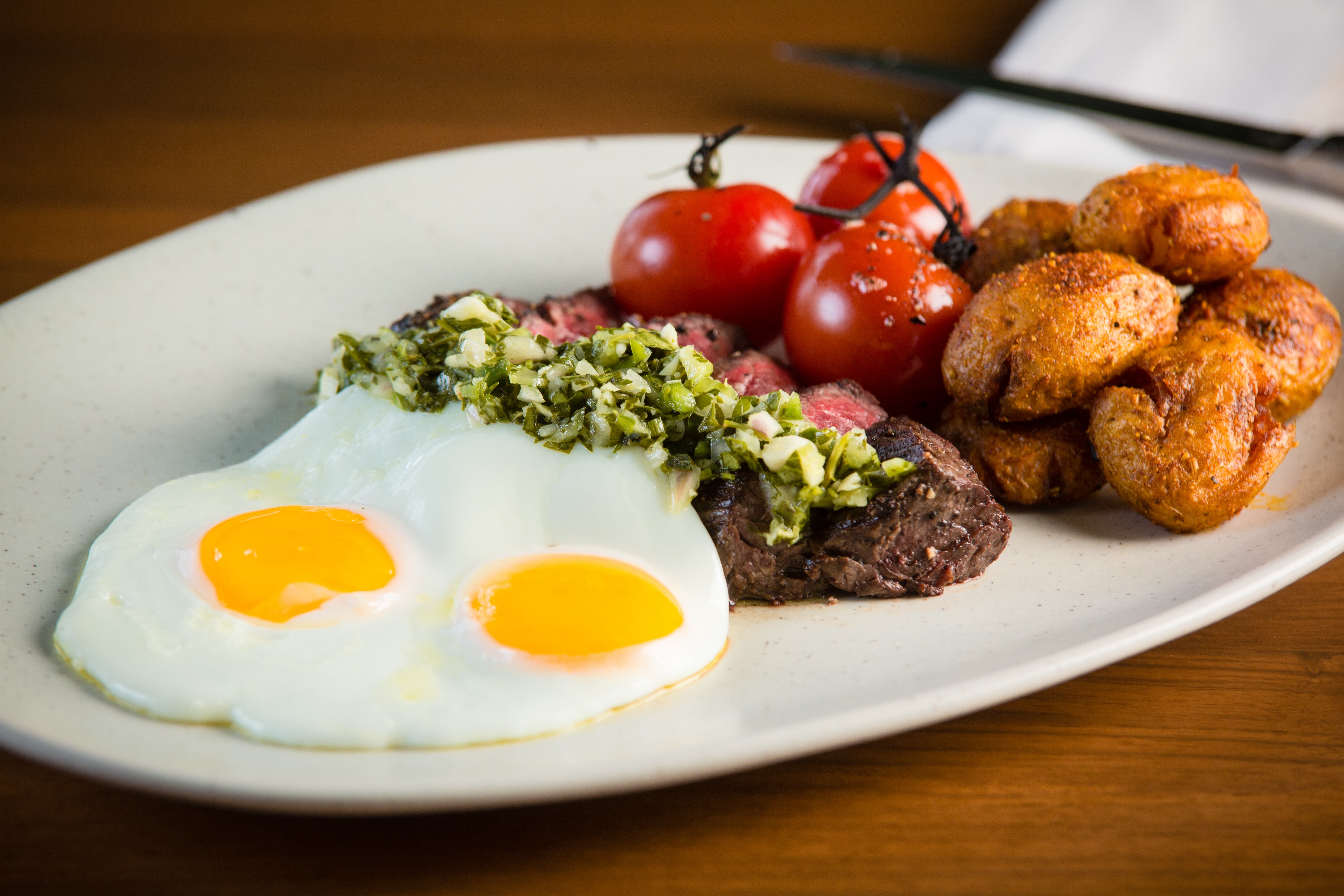 A perfect weekend brunch features a plate with two sunny-side-up eggs, grilled tomatoes, seasoned potatoes, and a steak topped with a green herb mixture.