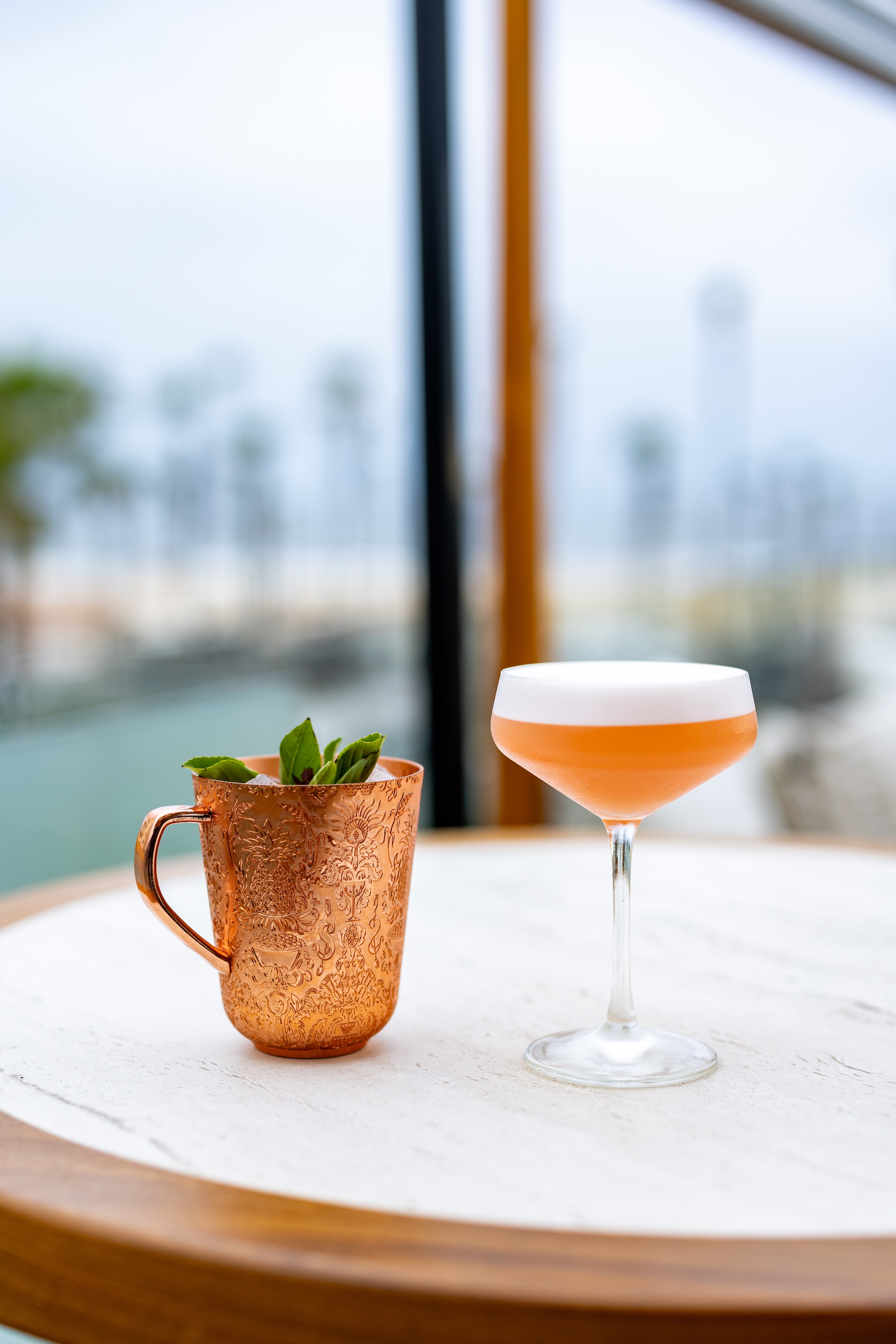 A copper mug filled with a mint garnish and a frothy cocktail in a coupe glass sit on a round marble table, capturing the essence of Happy Hour in the blurred outdoor background.
