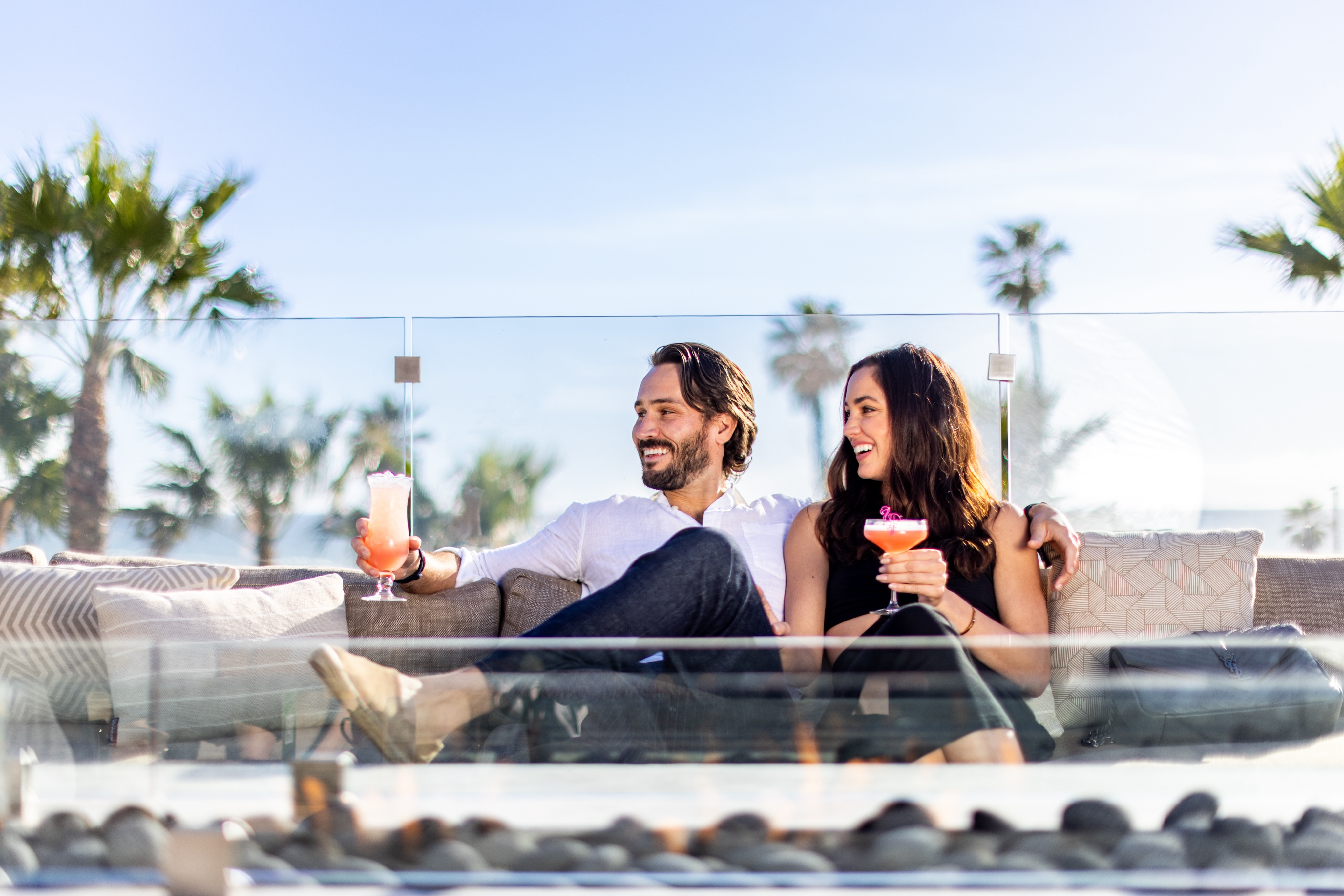 A man and a woman sit on an outdoor sofa, holding drinks, enjoying Happy Hour against the backdrop of palm trees and a glass barrier.