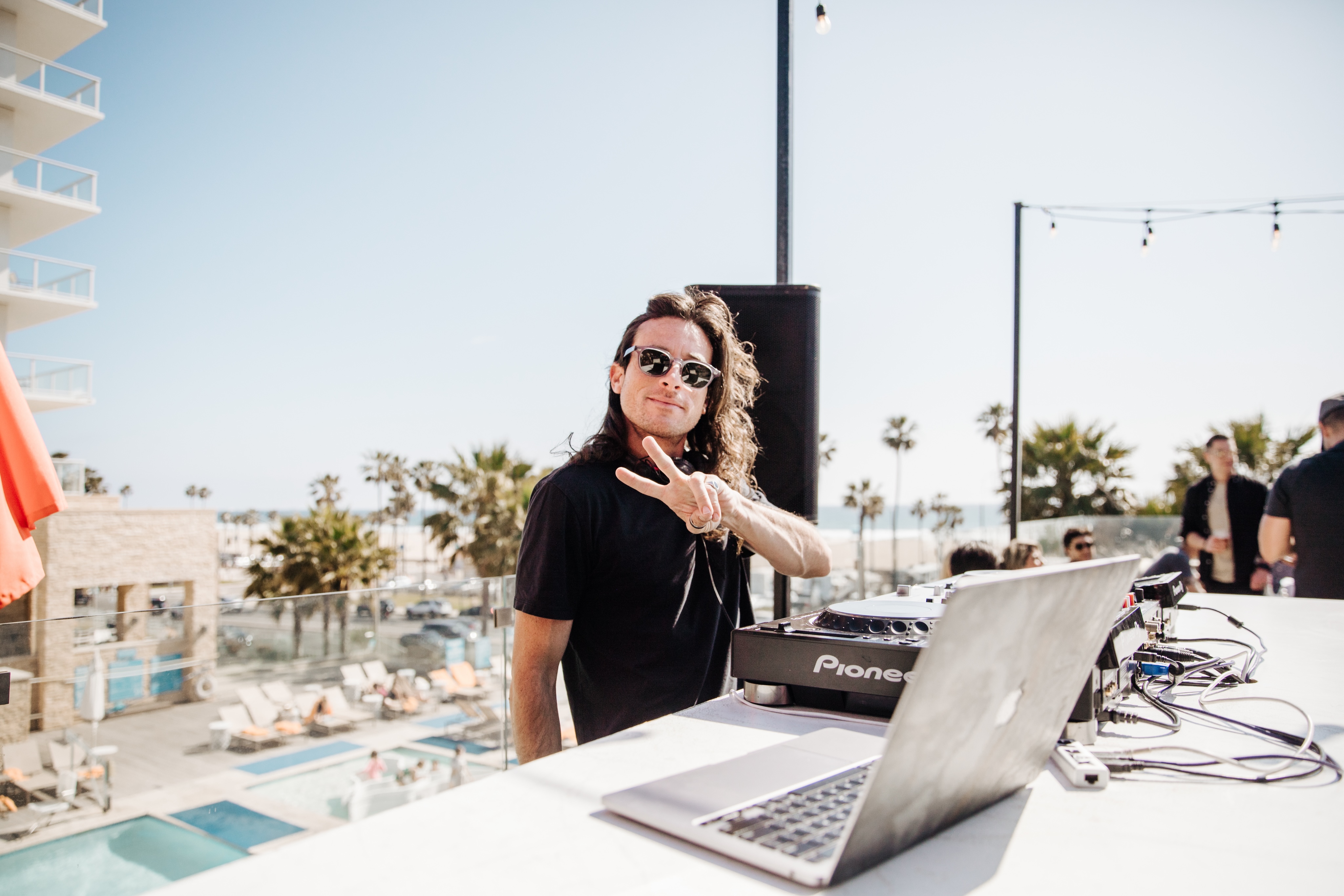 A DJ wearing sunglasses and a black shirt makes a peace sign while mixing music outdoors by a pool, delivering top-notch DJ entertainment at Treehouse on PCH.