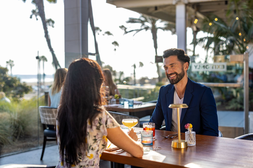 A man and a woman sit across from each other at an outdoor Lōrea restaurant table, engaged in conversation with drinks in front of them during Happy Hour. Palm trees and sunlight are visible in the background.