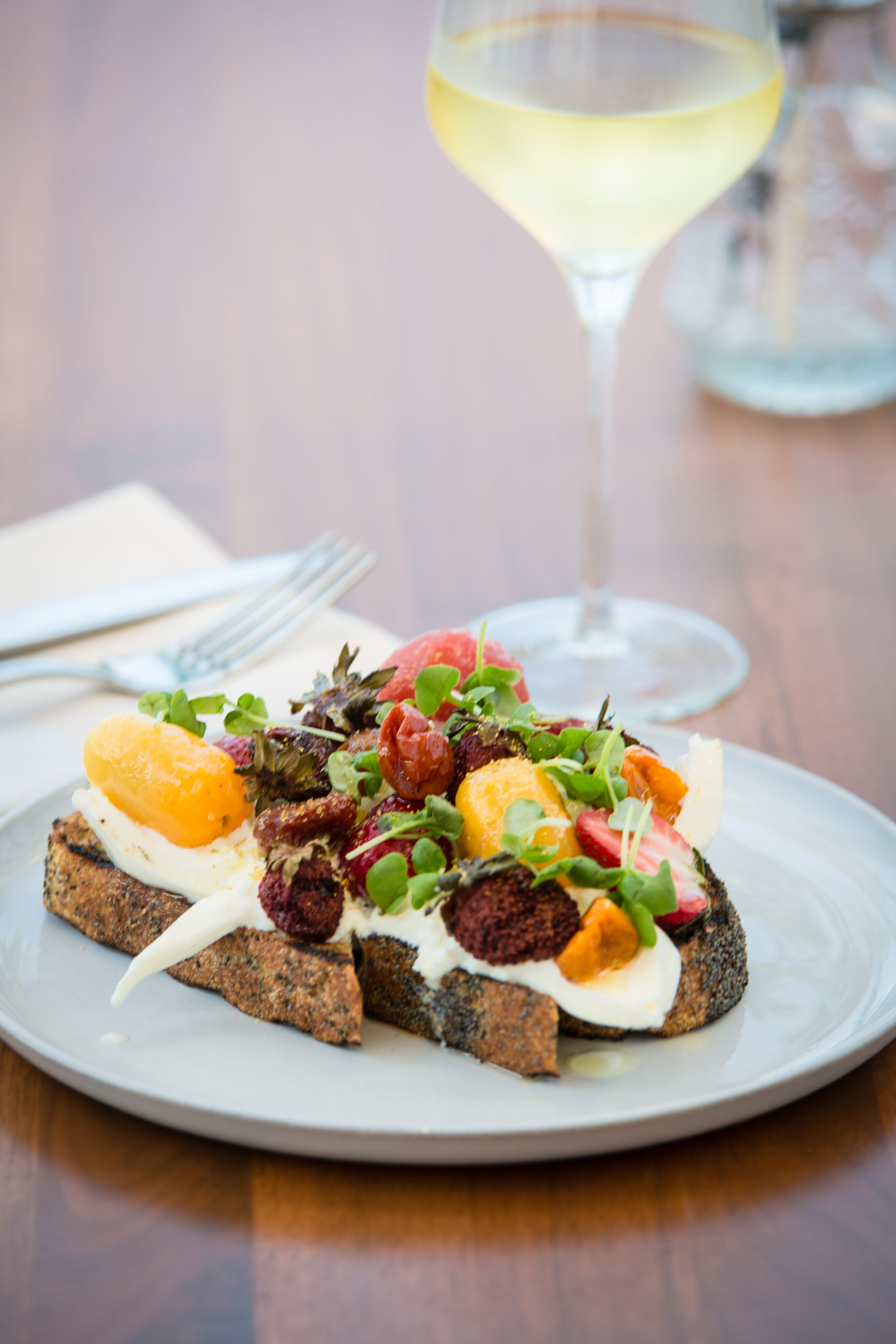 A plate of gourmet toast with various colorful toppings sits alongside a glass of white wine on a wooden table, creating an Instagram-worthy auto draft of culinary delight.