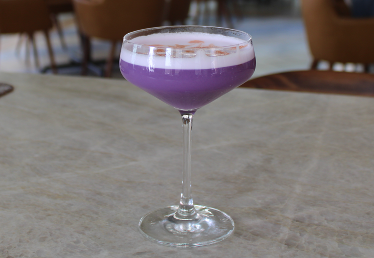 A purple cocktail in a stemmed glass with a white foamy top, garnished with a sprinkle of spice, sits mysteriously on the marble table, perfect for the Spooky Specials.