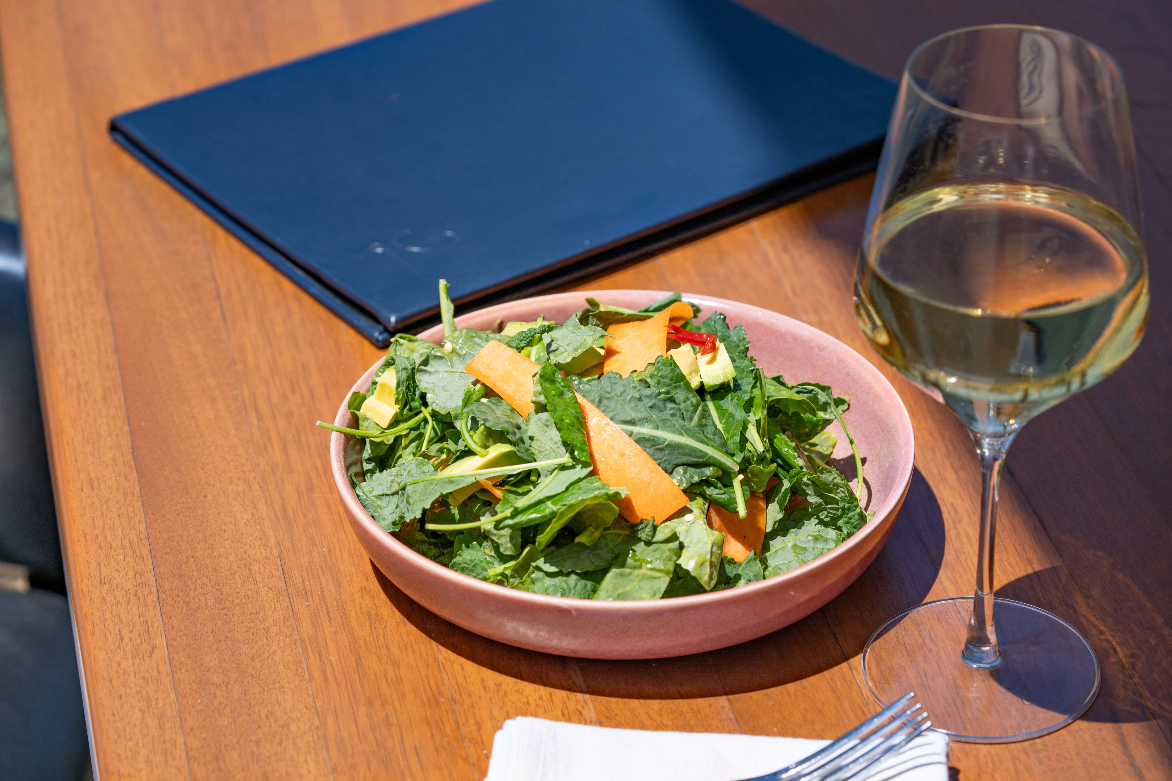 A bowl of green salad with sliced vegetables sits on a wooden table at Lōrea, perfect for a Power Lunch, next to a glass of white wine, a black menu, and a folded napkin with cutlery.