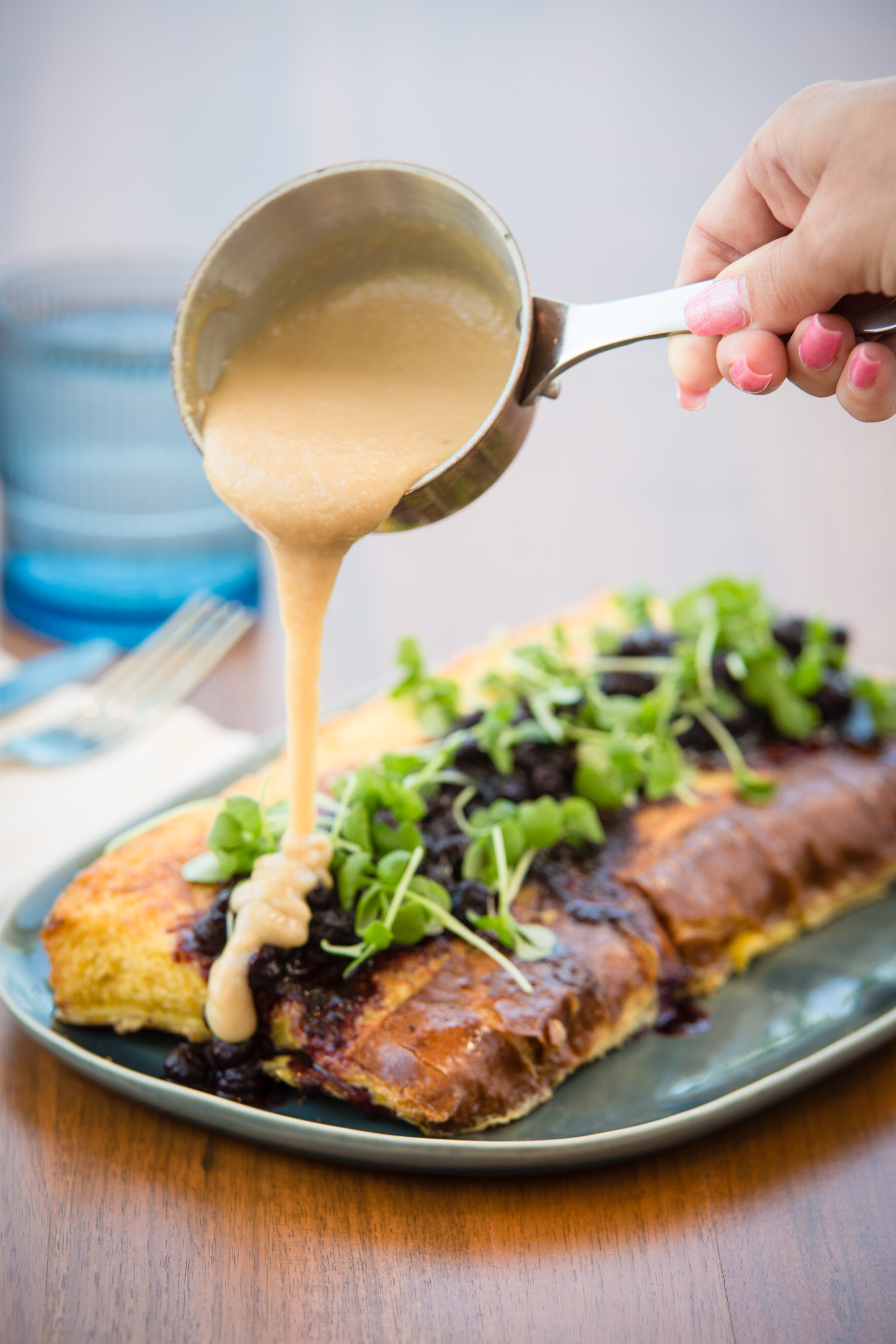 A hand pouring sauce over a large baguette topped with greens and a dark filling on a blue plate, perfect for curing that New Year's Day hangover.