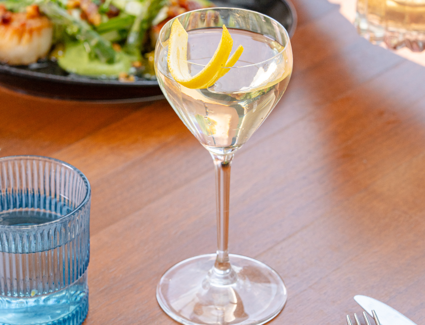 A clear cocktail from the Happy Hour Menu, garnished with a lemon twist, is served in a stemmed glass on a wooden table beside a blue water glass and a plate of food.