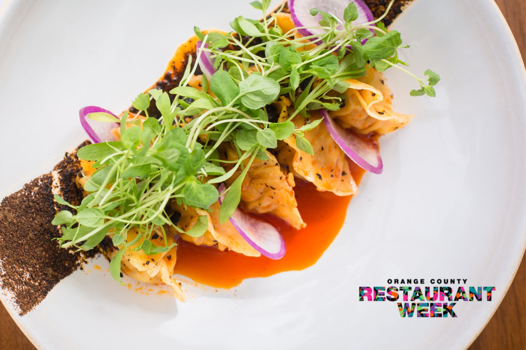 Four dumplings topped with microgreens and red onion slices, served with red sauce and spice on a white plate; featured for Orange County Restaurant Week—the logo appears in the corner.