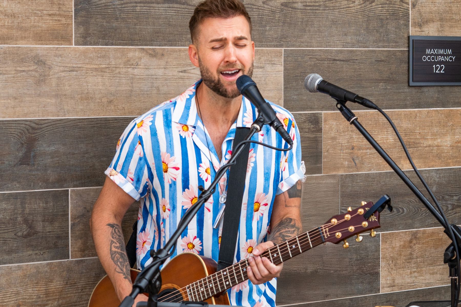 A man in a floral striped shirt plays an acoustic guitar and sings into a microphone for the Lōrea Music Series in an outdoor setting.