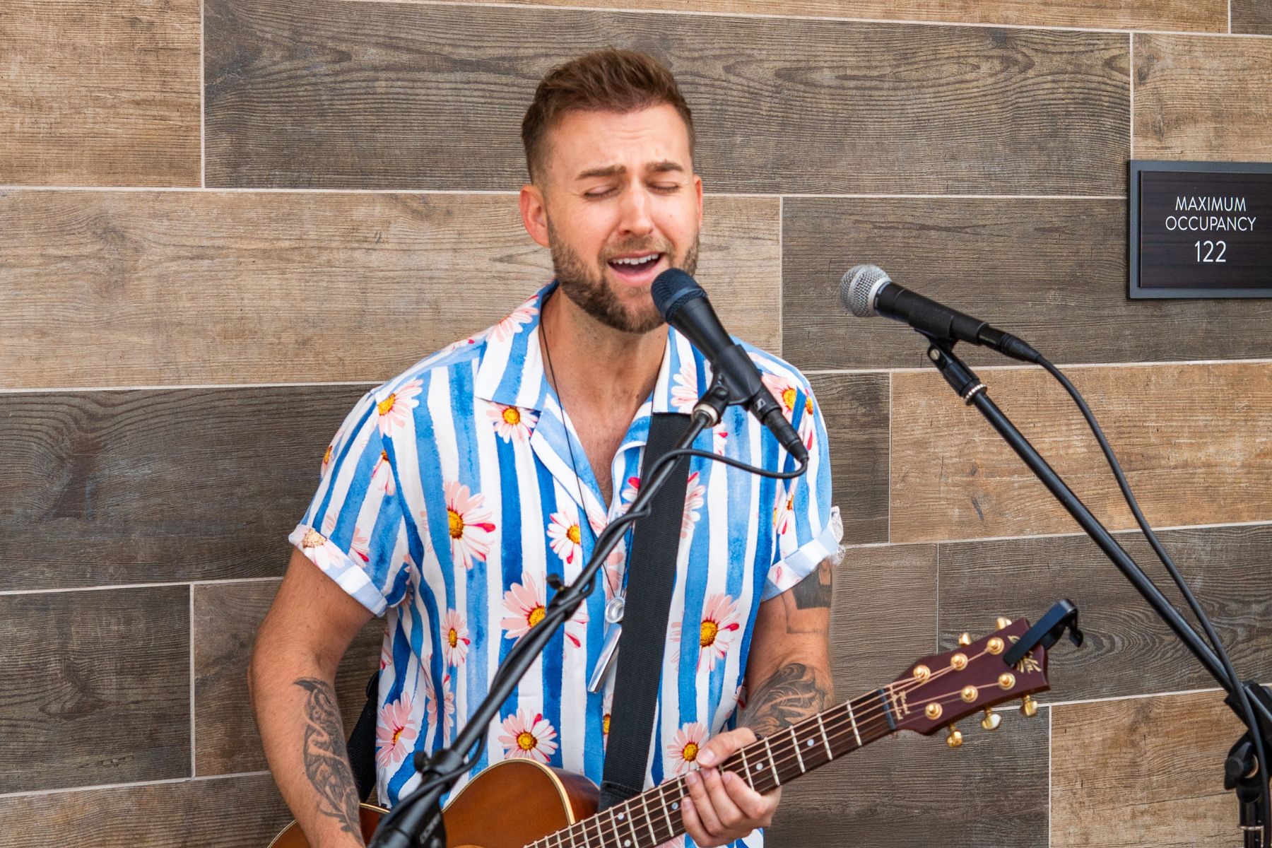 A man wearing a floral, striped shirt sings and plays an acoustic guitar into a microphone in front of a wood-paneled wall for the Lōrea Music Series.