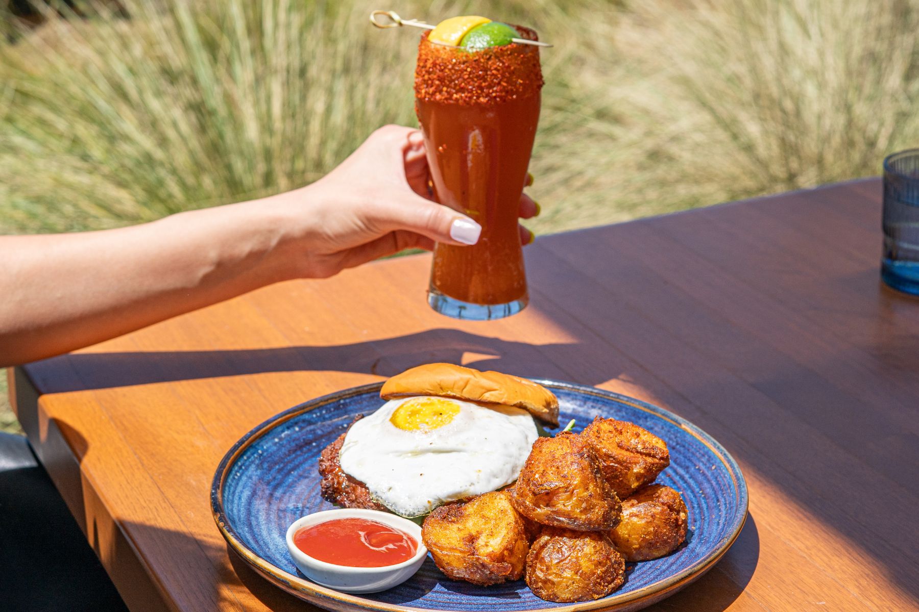 A person holds a glass of a red cocktail with a lime garnish above a plate featuring a fried egg burger, crispy potatoes, and ketchup—perfect for an Easter Brunch enjoyed outdoors on a wooden table.