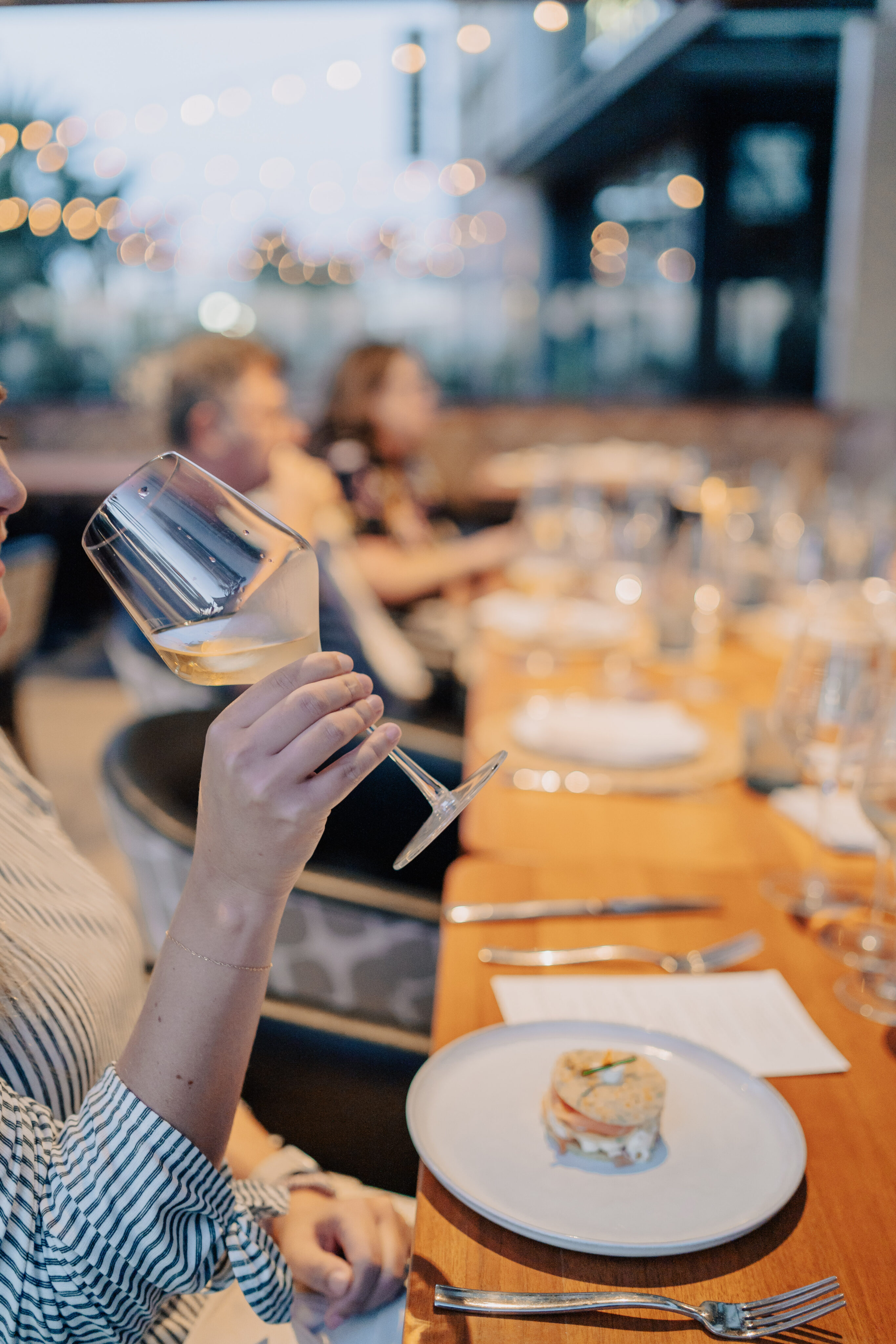 A person elegantly holds a wine glass at a dining table adorned with plates and cutlery, as whispers of soirees fill the air with blurred figures in the background and the gentle glow of ambient lighting, evoking a sense of springtime gatherings.