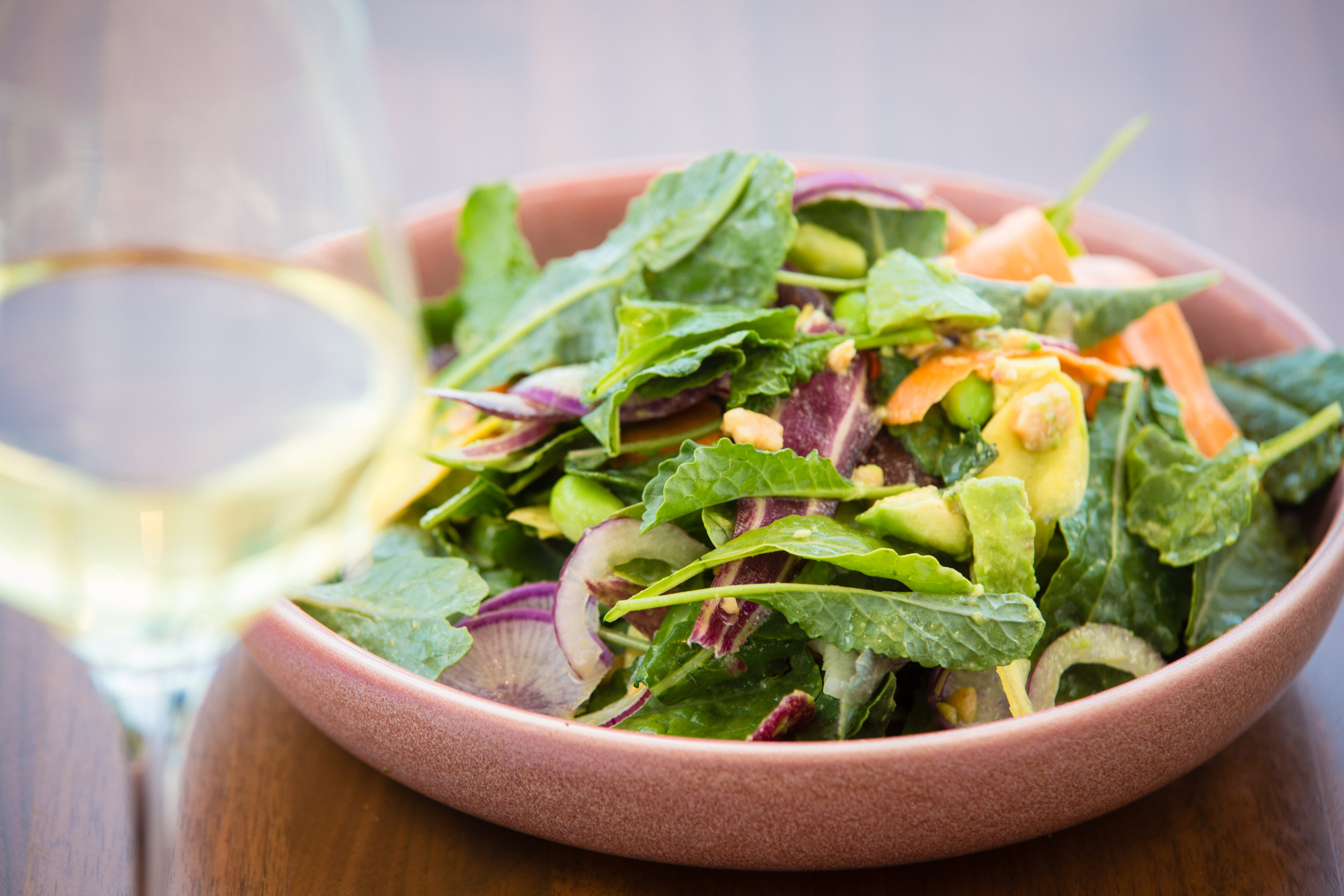 A bowl of mixed green salad with purple onions, avocado, and carrots beside a glass of white wine on a wooden table.