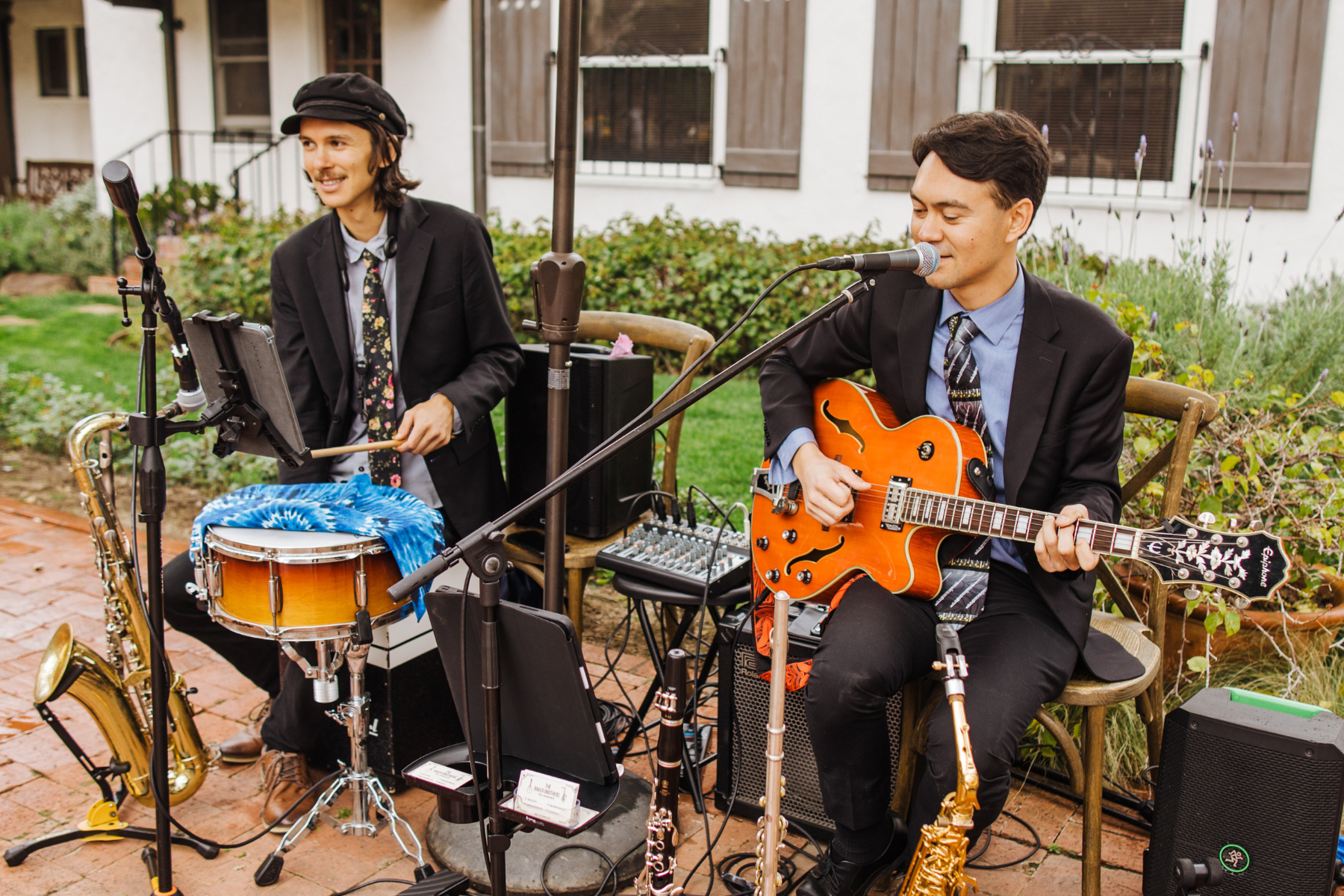 Two musicians perform outdoors at Jazz Night; one plays drums and the other, representing Lōrea, sings while playing an electric guitar. Various musical equipment and instruments are visible around them.