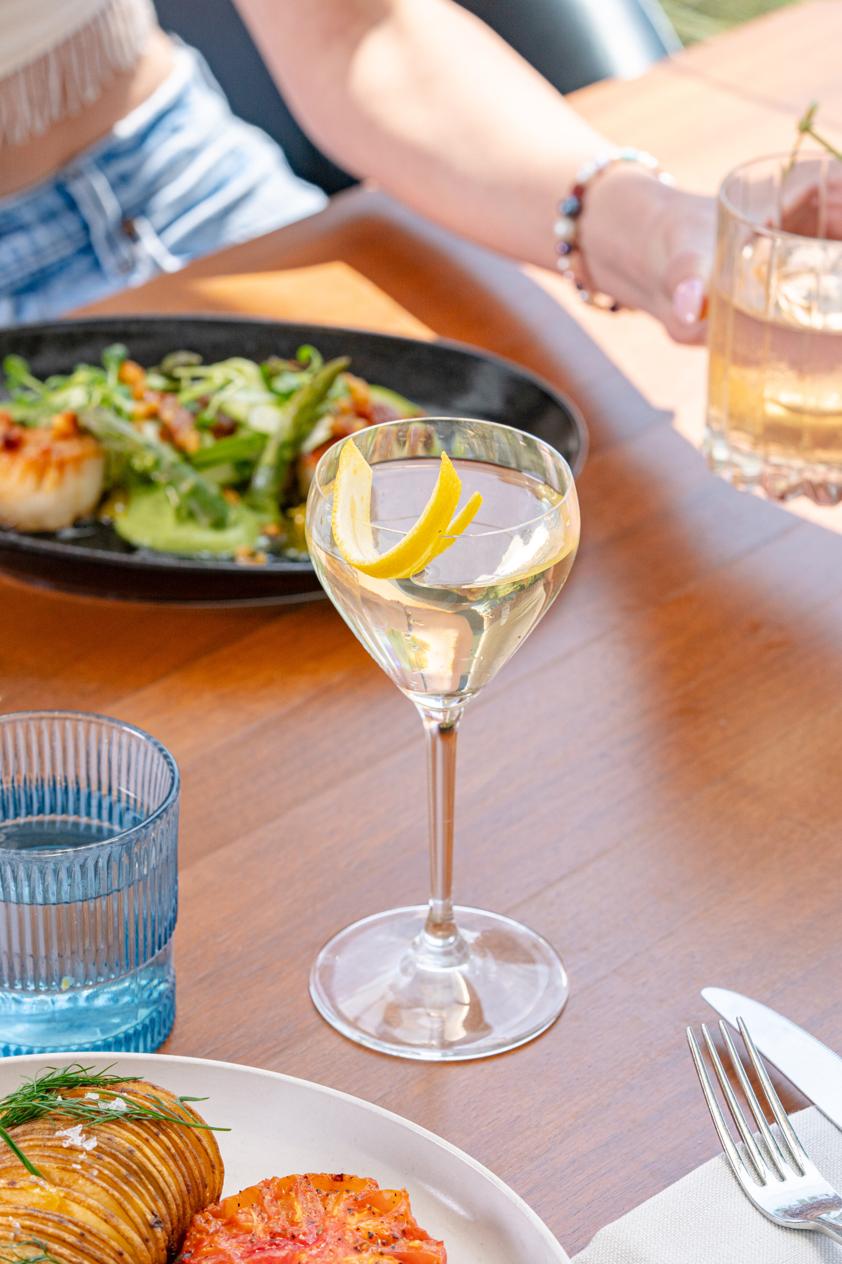 A close-up of a table at Lōrea during Jazz Night shows a clear cocktail garnished with lemon peel, a blue glass of water, plates of food, and a person reaching for a drink.