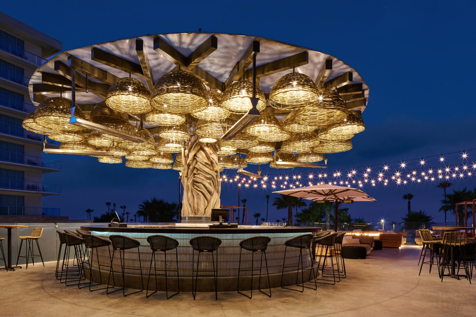 Circular outdoor bar with a central treehouse-inspired structure supporting woven pendant lights, surrounded by barstools—perfect for an Agave After Dark Party—with string lights and palm trees glowing in the dusk.