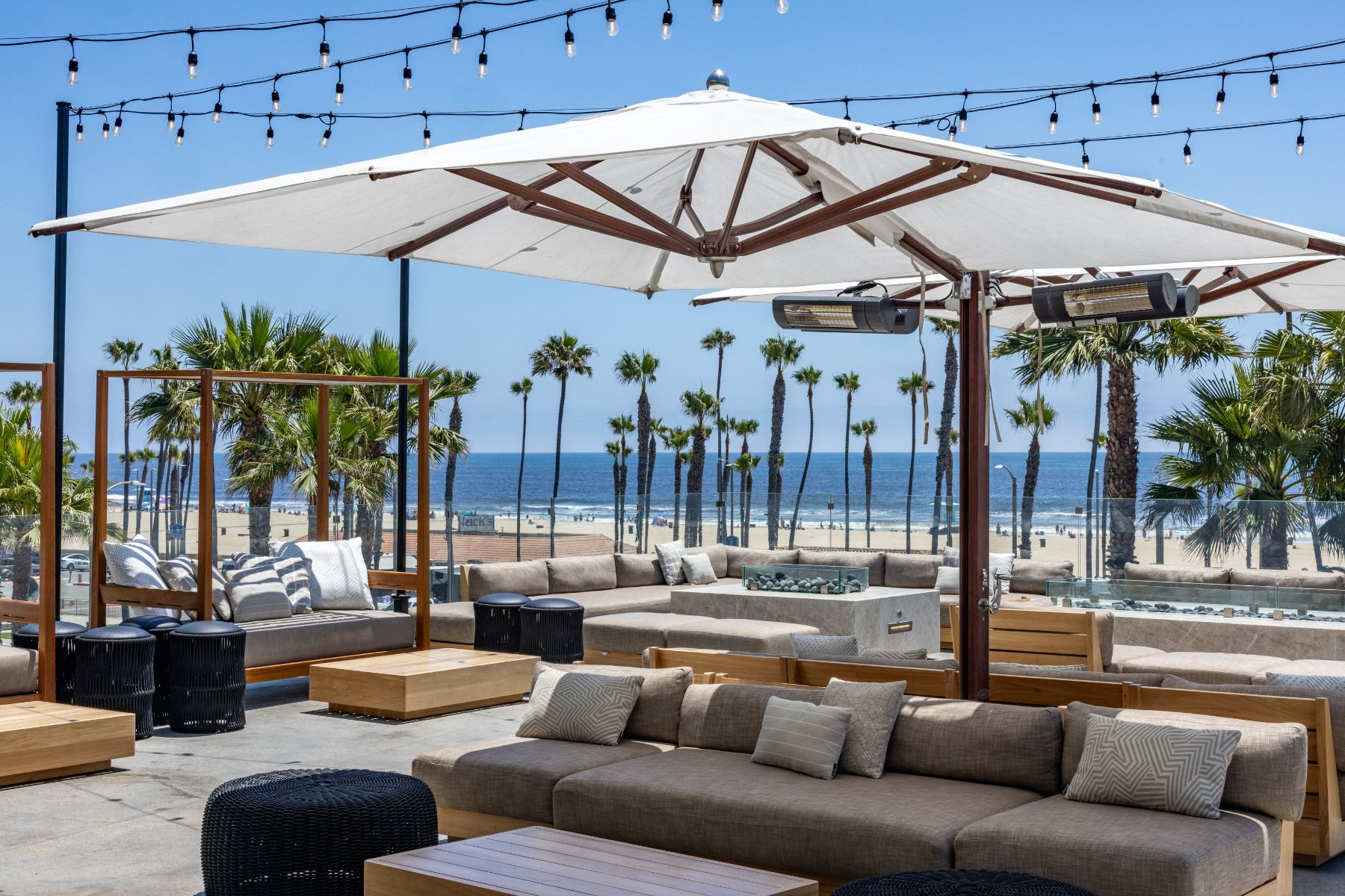 Outdoor patio with beige couches, wooden tables, large umbrellas, and string lights exudes a cozy Treehouse vibe, surrounded by palm trees and overlooking a sandy beach and ocean under a clear blue sky.
