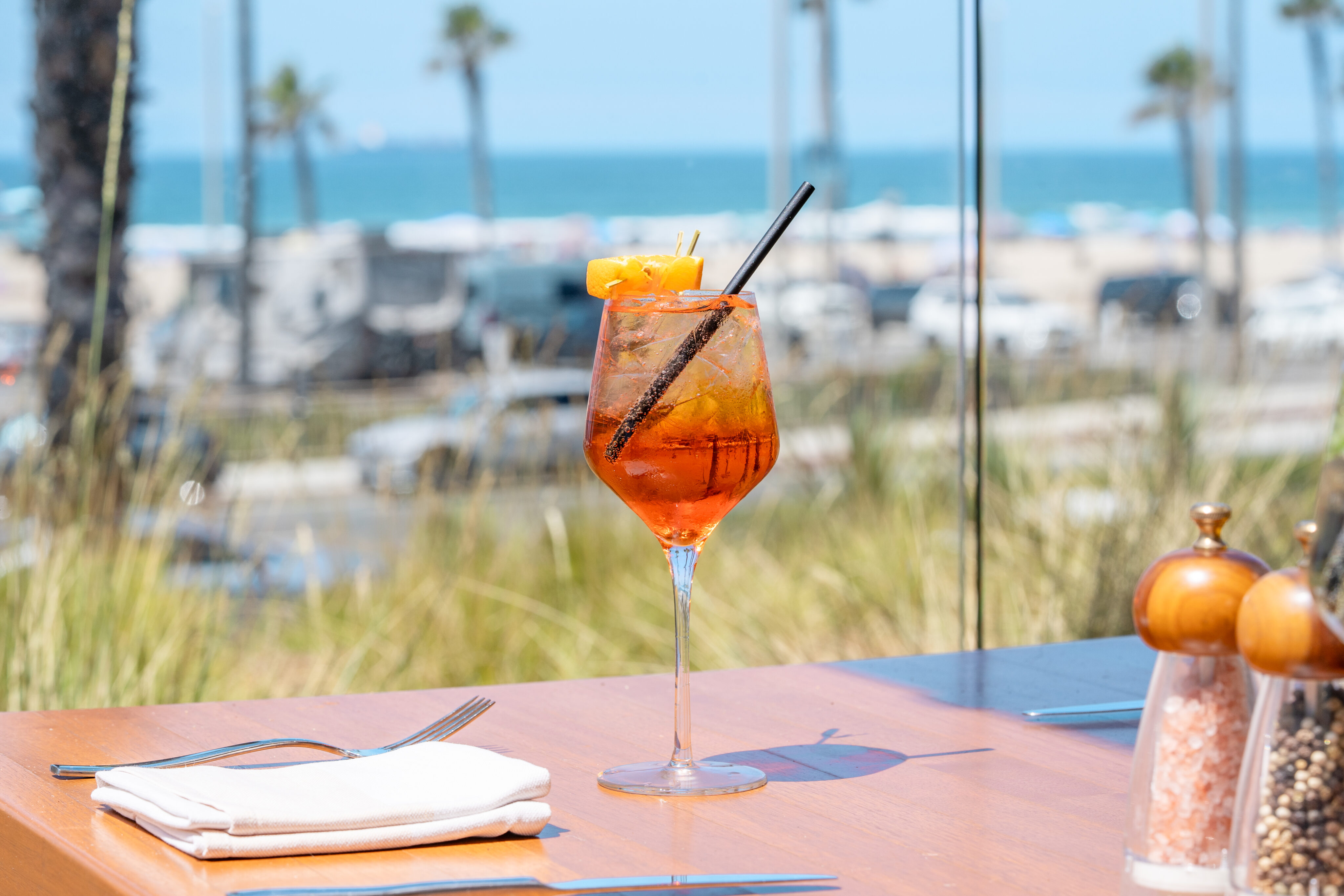 A glass of orange Spritz with ice and orange garnish sits on a wooden table set with cutlery, napkin, salt, and pepper—perfect for a Weekend Brunch with a stunning beach and ocean view in the background.