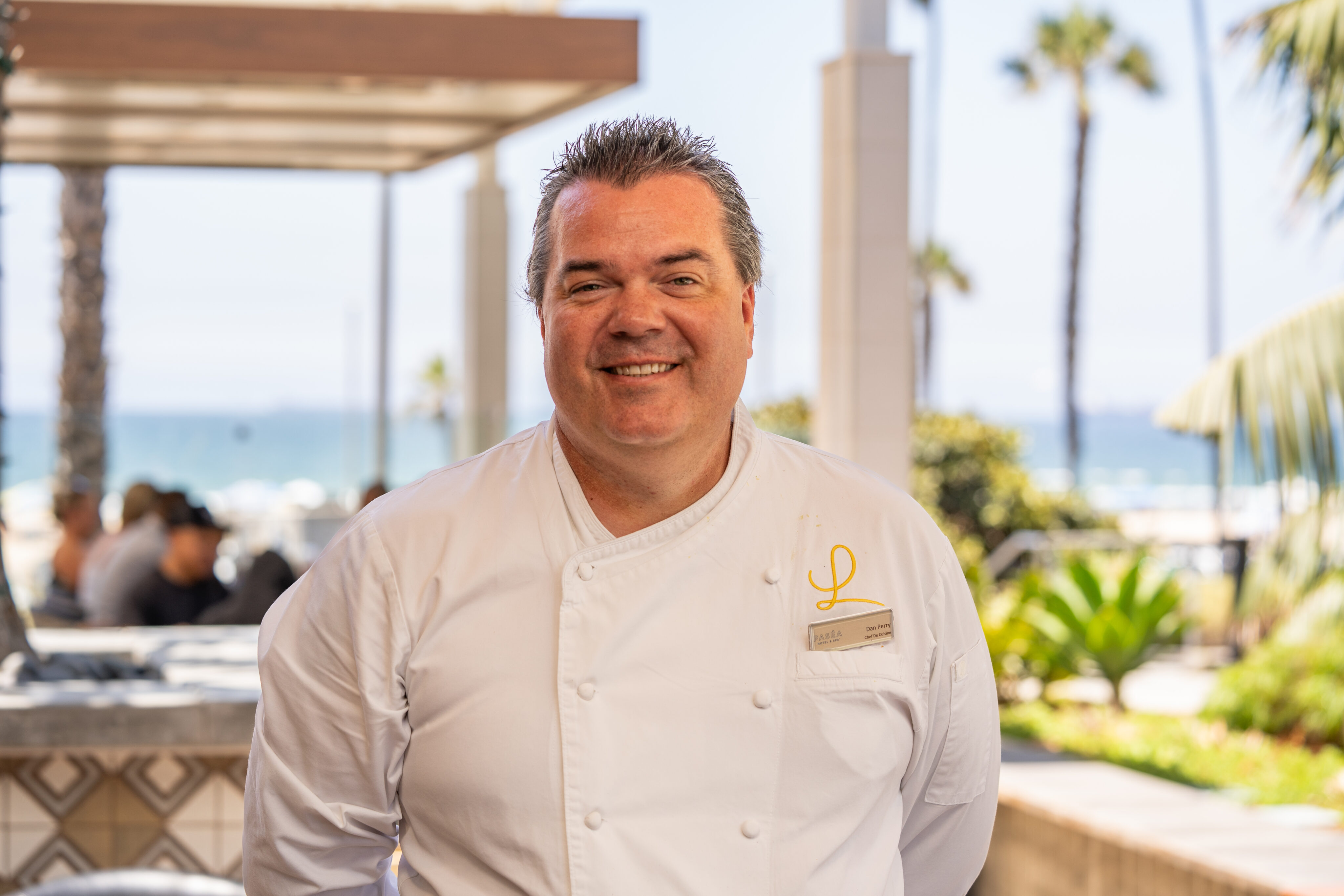 A chef in a white uniform stands outdoors near a beach, smiling at the camera with palm trees and the ocean in the background.