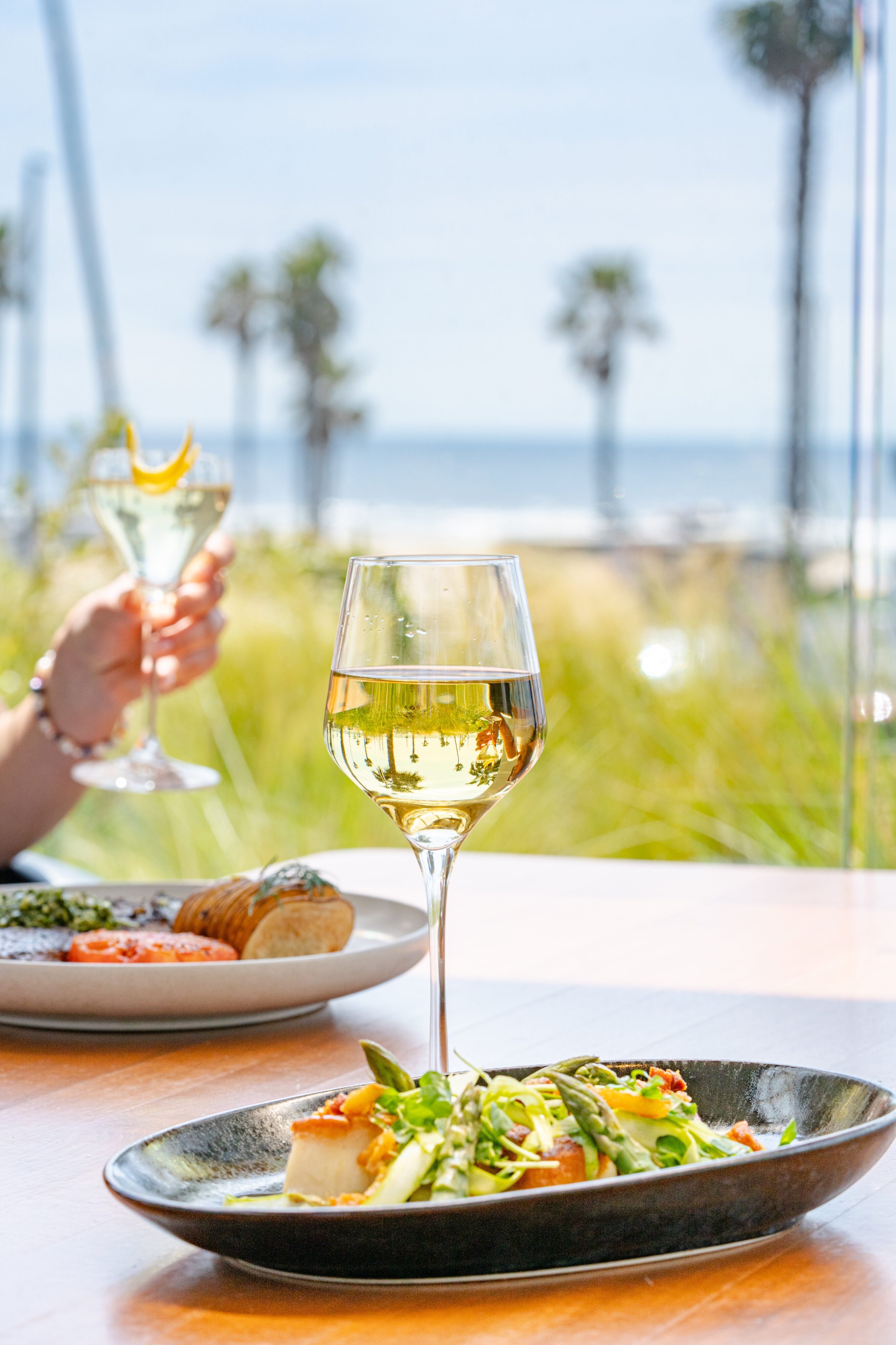 A glass of white wine and a plate of food sit on a table by a window at Lorea Dining, with a blurred beach and palm trees visible in the background.