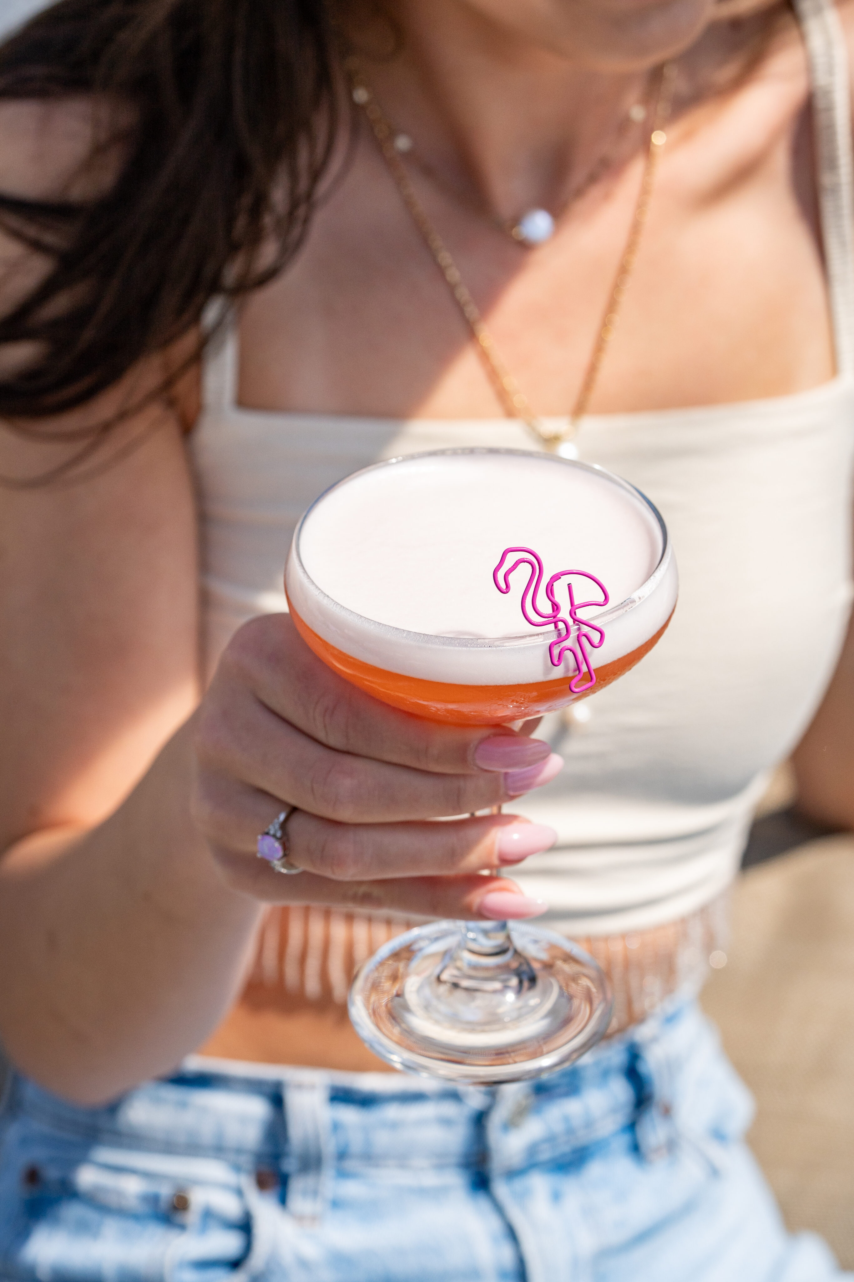 A woman in a white crop top and blue jeans enjoys a pink cocktail topped with foam and a pink flamingo stir stick during Tequila Tuesdays.