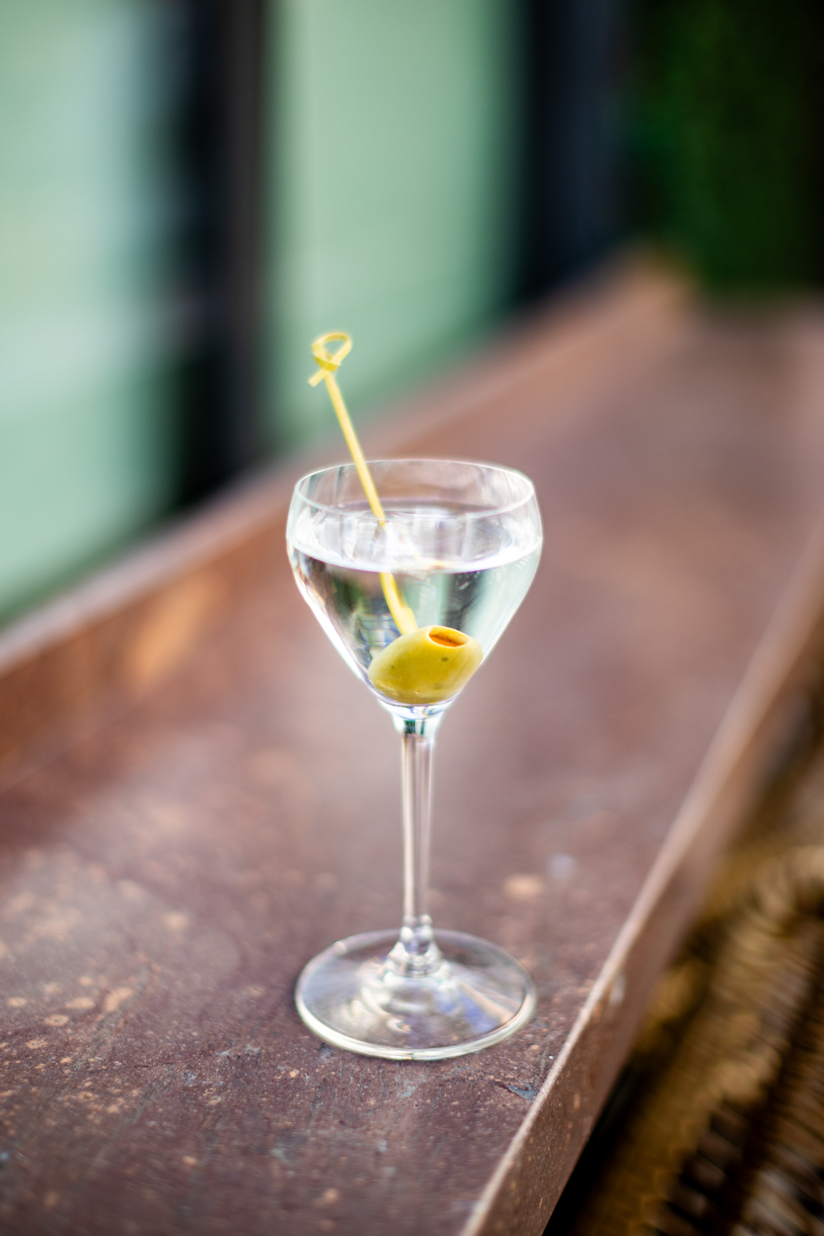 A clear cocktail served in a stemmed glass with a single green olive on a cocktail pick, set on a brown surface—perfect for savoring at Treehouse during Weber Wednesdays.