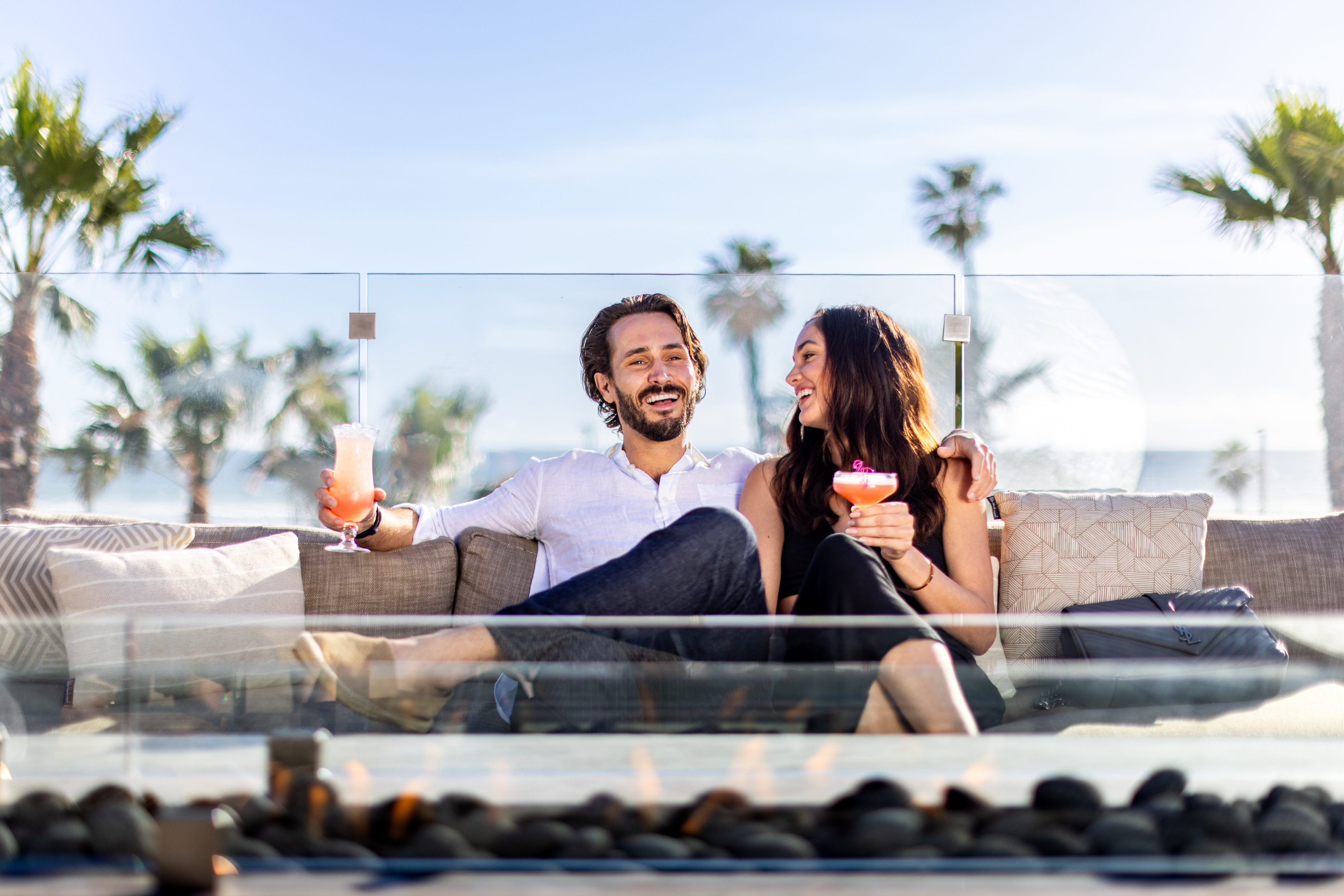 A man and woman sit on an outdoor sofa at Treehouse, holding cocktails and smiling at each other, with palm trees and a clear sky in the background—perfect vibes for Tequila Tuesdays.