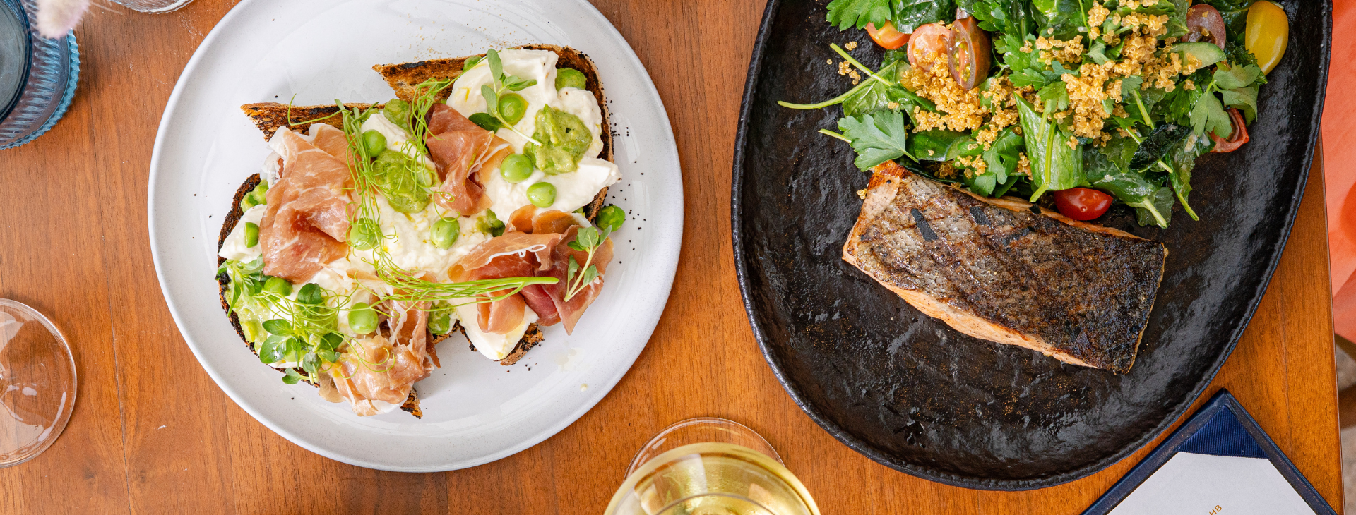 A plate with toast topped with prosciutto, mozzarella, and greens sits beside a plate with grilled salmon and salad at Lorea Dining on a rustic wooden table.