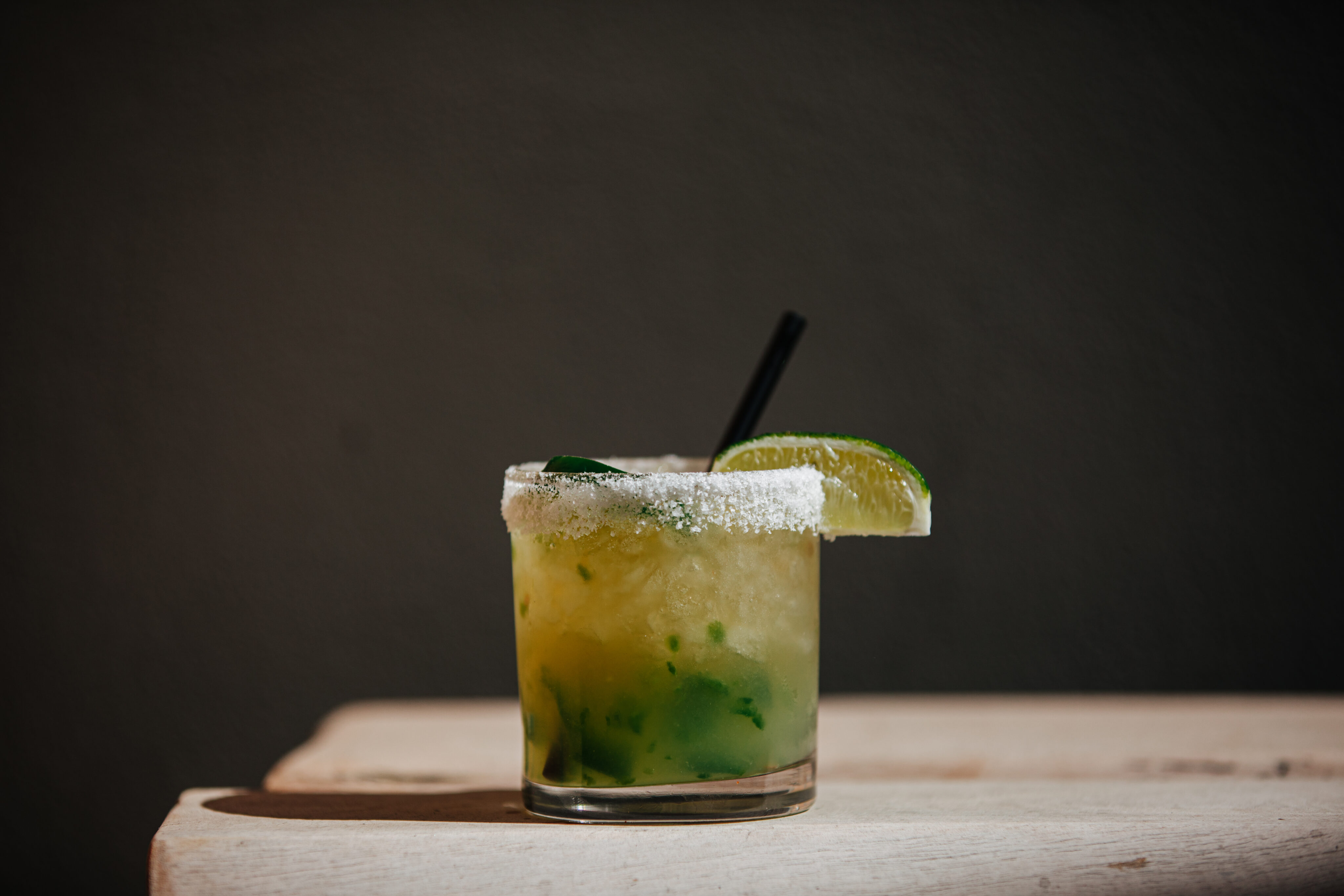 A salt-rimmed glass filled with a yellow-green Tequila cocktail, ice, mint leaves, and a lime wedge, with a black straw, sits on a light wooden surface against a dark background—perfect for celebrating National Tequila Day at Treehouse.