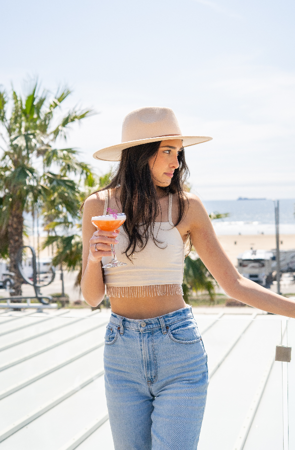 A woman in a hat and crop top holds a cocktail made with Código 1530 Tequila while standing outdoors by a railing, with a beach and palm trees in the background.