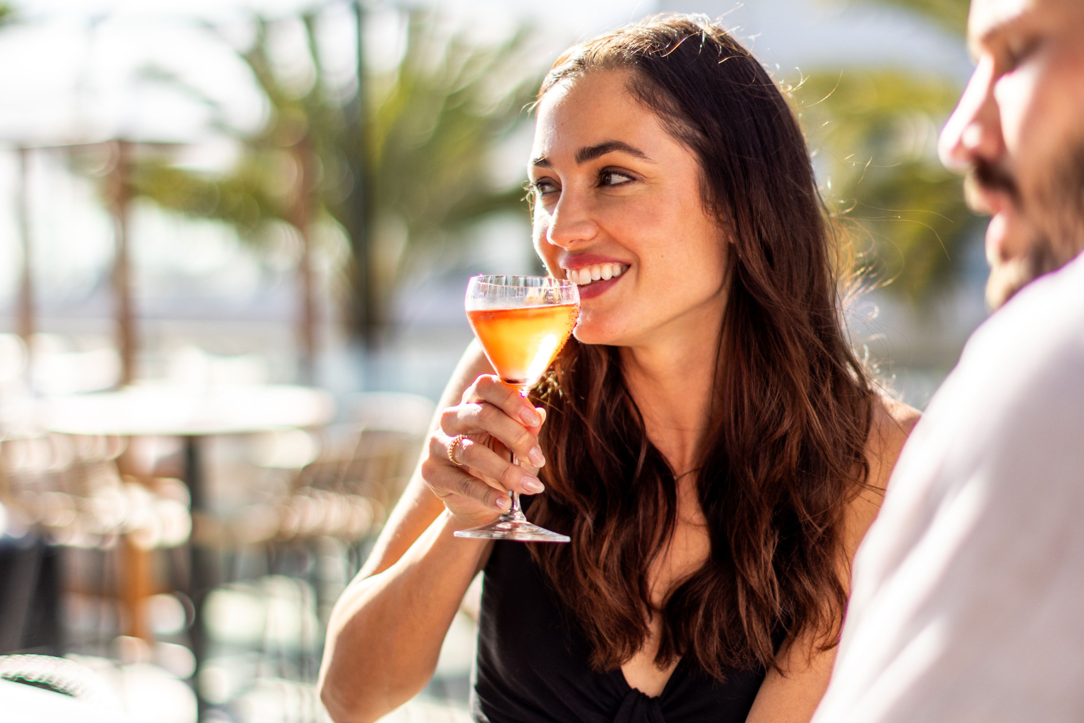 A woman with long brown hair smiles while holding a cocktail glass at Treehouse on PCH, sitting outdoors next to a man, with blurred palm trees in the background—perfect for enjoying Spooky Specials.