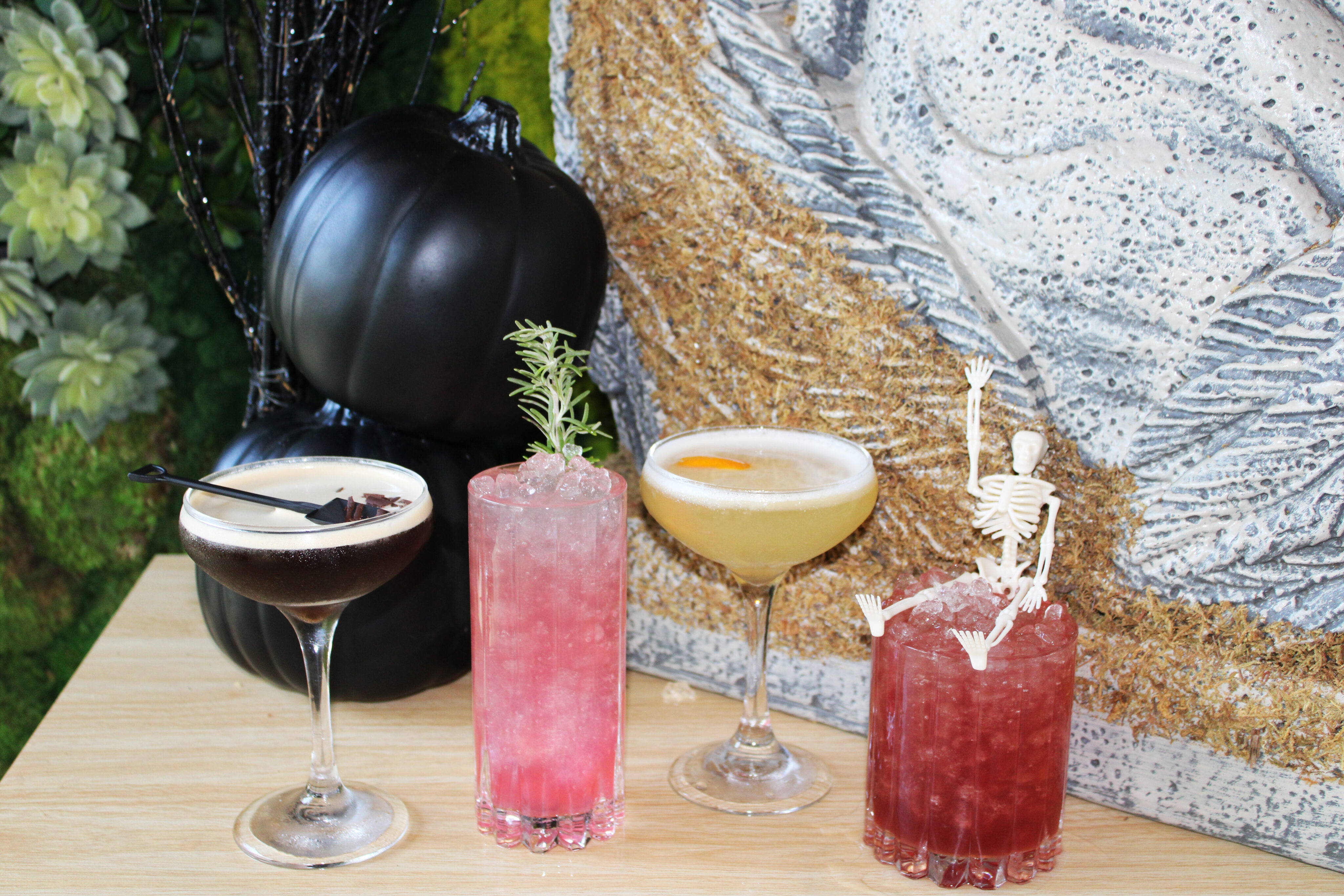 Four cocktails on a wooden table—two with garnishes, one featuring a plastic skeleton—set against black pumpkins and textured artwork. Enjoy these Spooky Specials at Treehouse, perfect for a haunting night out near PCH.