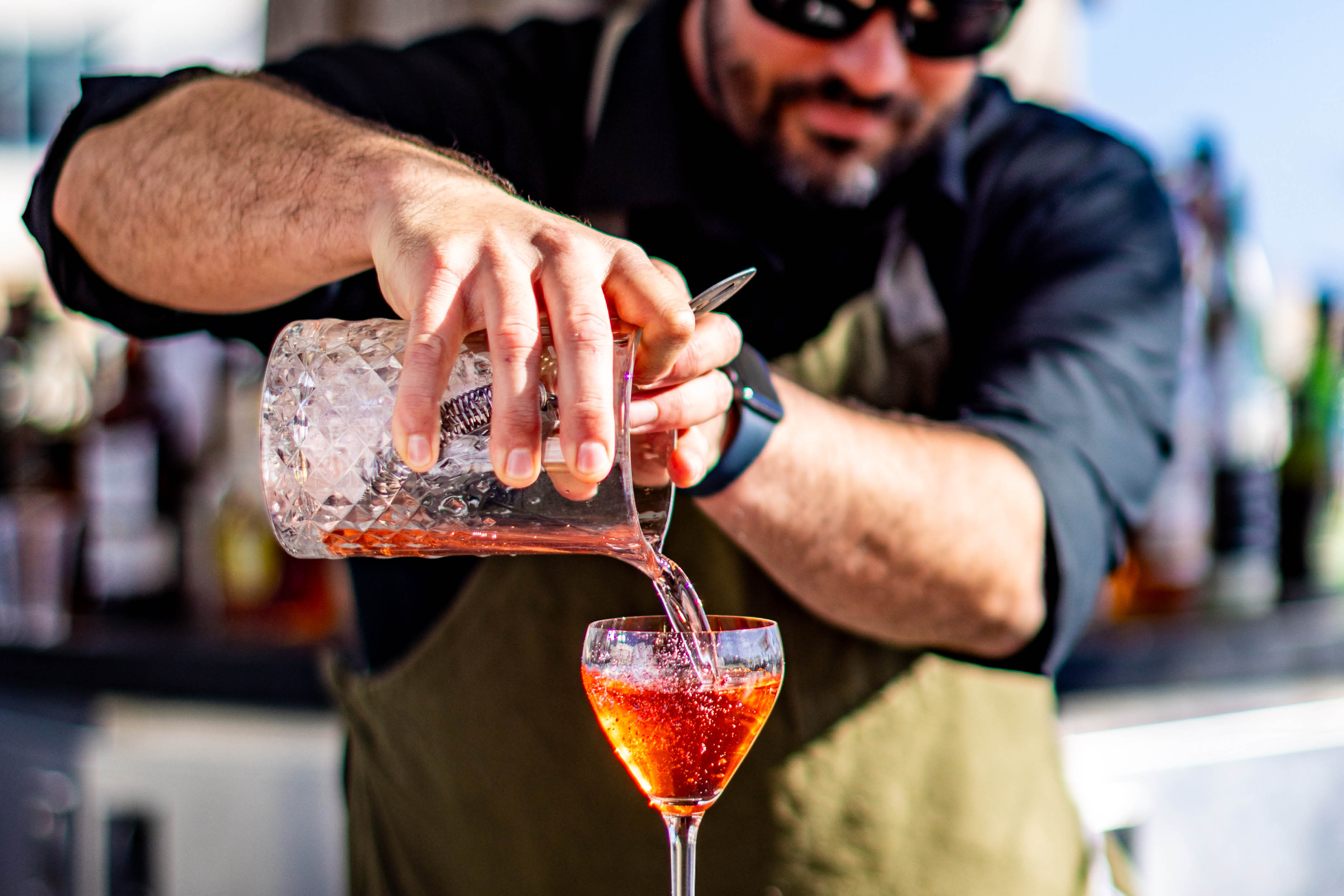 A bartender in sunglasses and an apron pours a red cocktail from a mixing glass into a stemmed glass at Treehouse during Industry Nights.