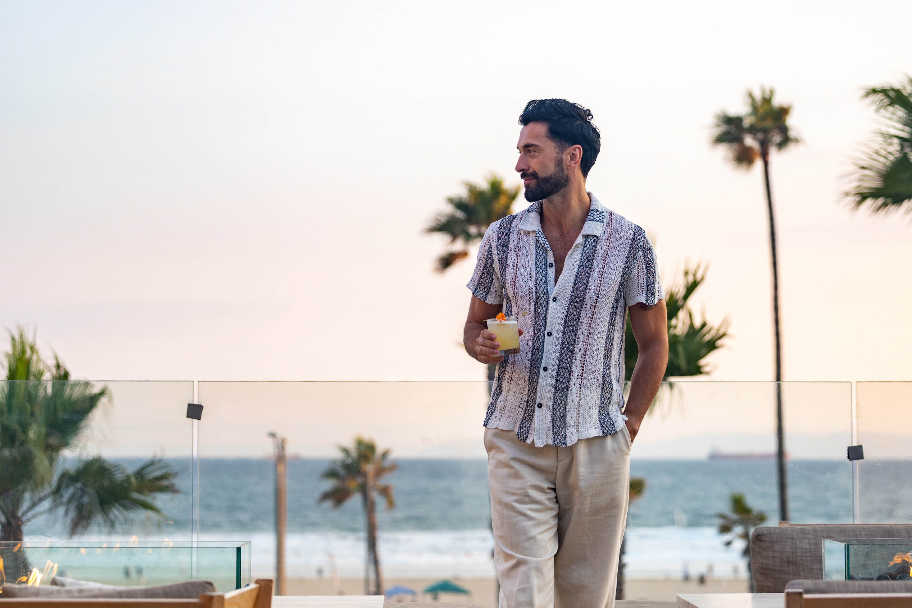 A man holding a drink strolls on the Treehouse terrace, overlooking a beach with palm trees during Sunset Sessions.