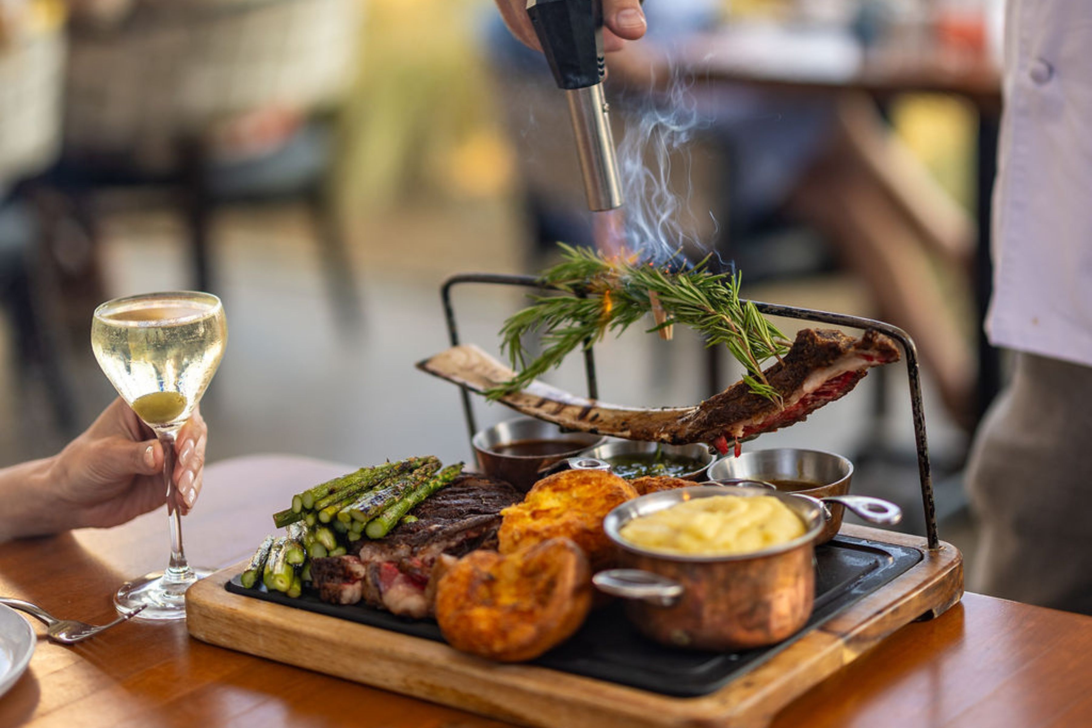 A chef uses a blowtorch on a rib steak garnished with herbs, served with asparagus, potatoes, and a glass of white wine at a restaurant table.