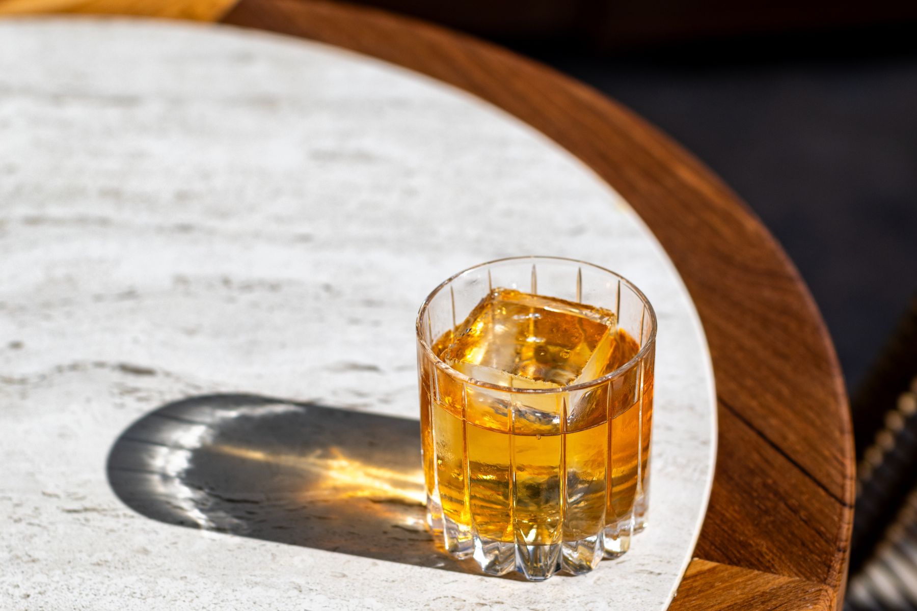A glass of amber-colored liquor with ice sits on a white and wooden tabletop, casting a shadow in sunlight.