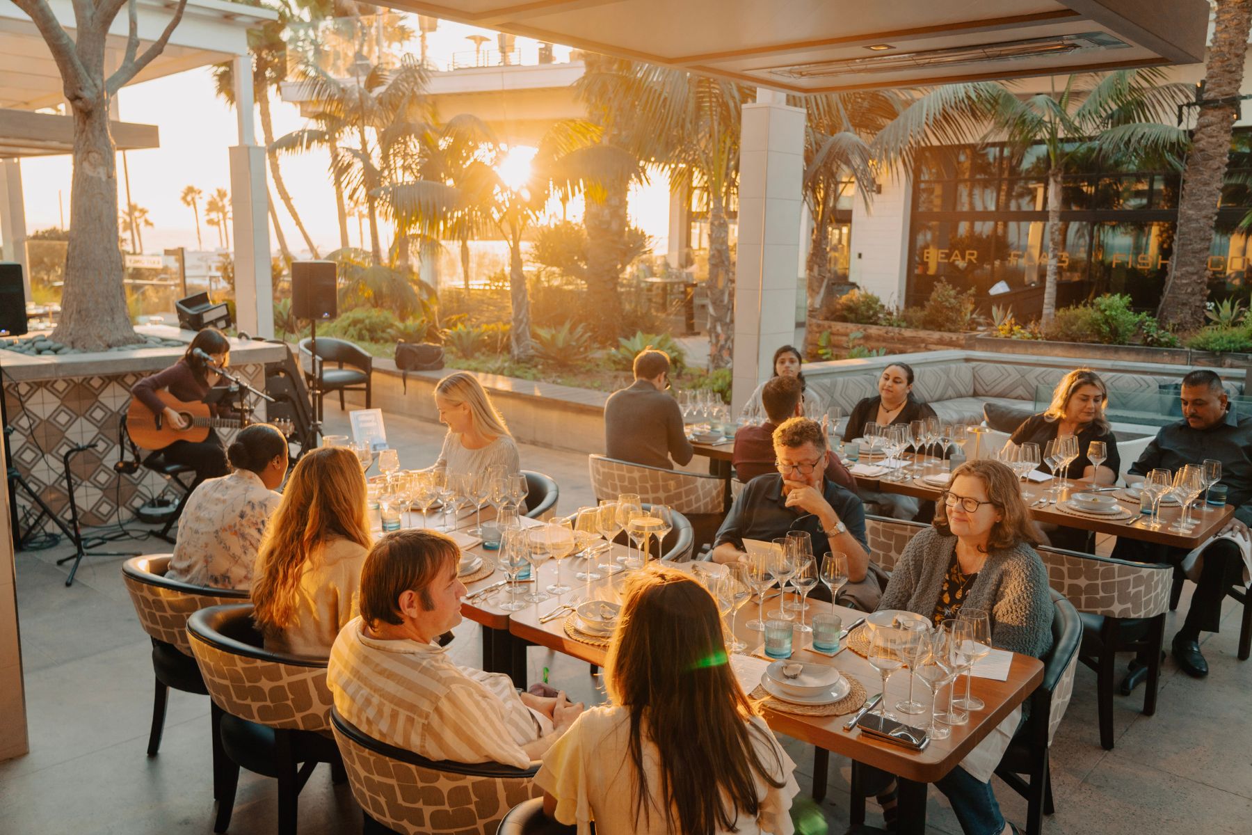 People enjoying Holiday Private Dining at an outdoor restaurant during sunset, with a musician playing guitar and palm trees surrounding the patio.
